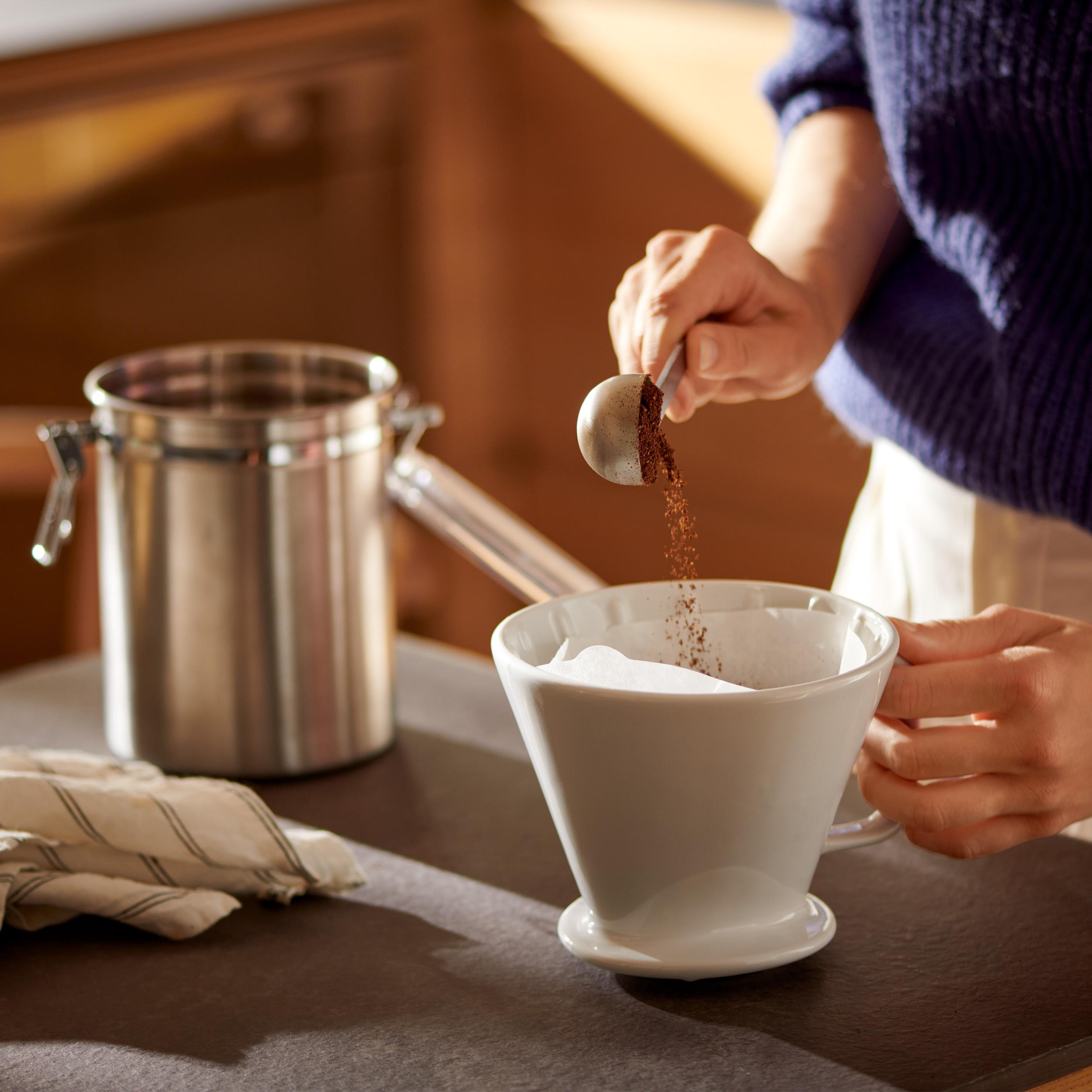 Eine Person gießt Kaffeepulver in einen weißen Filter in einem weißen Keramik-Pour-Over-Kaffeebereiter.
