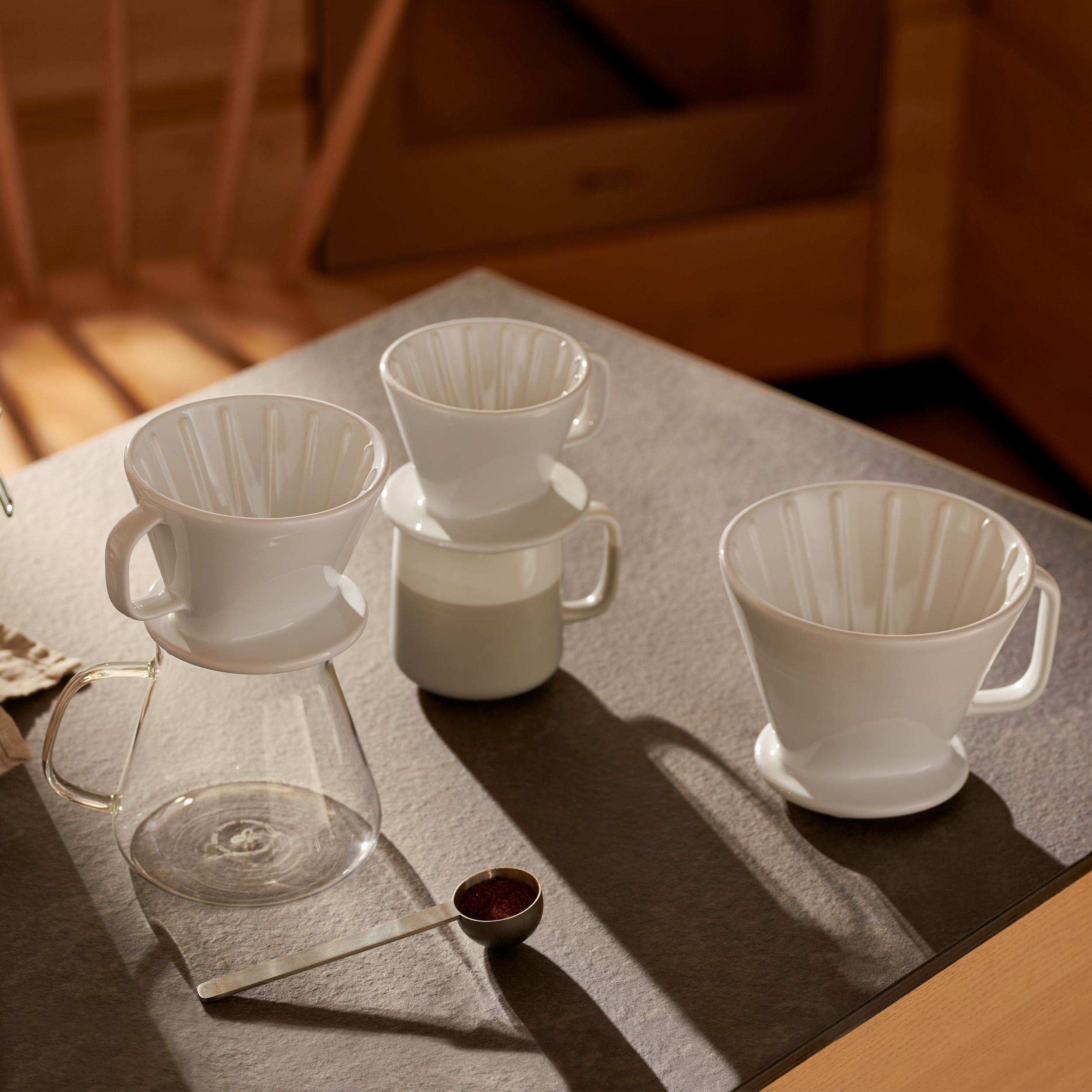 Drei weiße Pour-Over-Kaffeebereiter auf einem Tisch, mit einer Kaffeepulver-Schaufel in der Nähe.