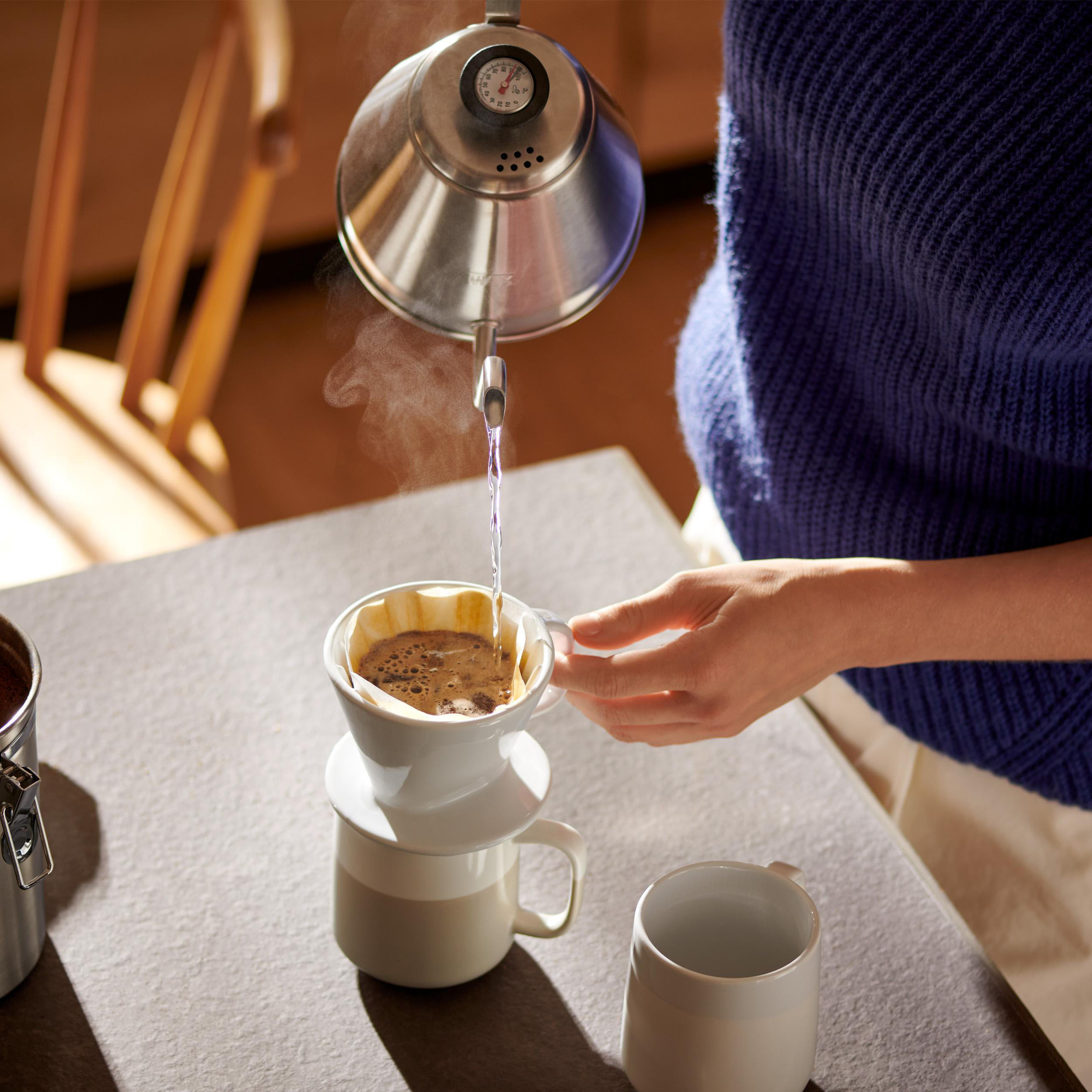 Person gießt heißes Wasser in einen Kaffeefilter, Keramikbecher in der Nähe.