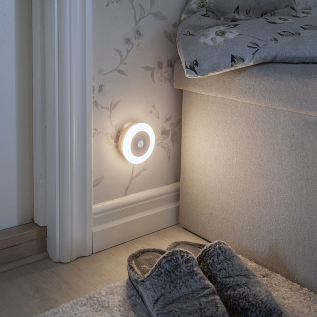 Luce notturna a parete, luminosa, accanto a un letto e a delle pantofole.