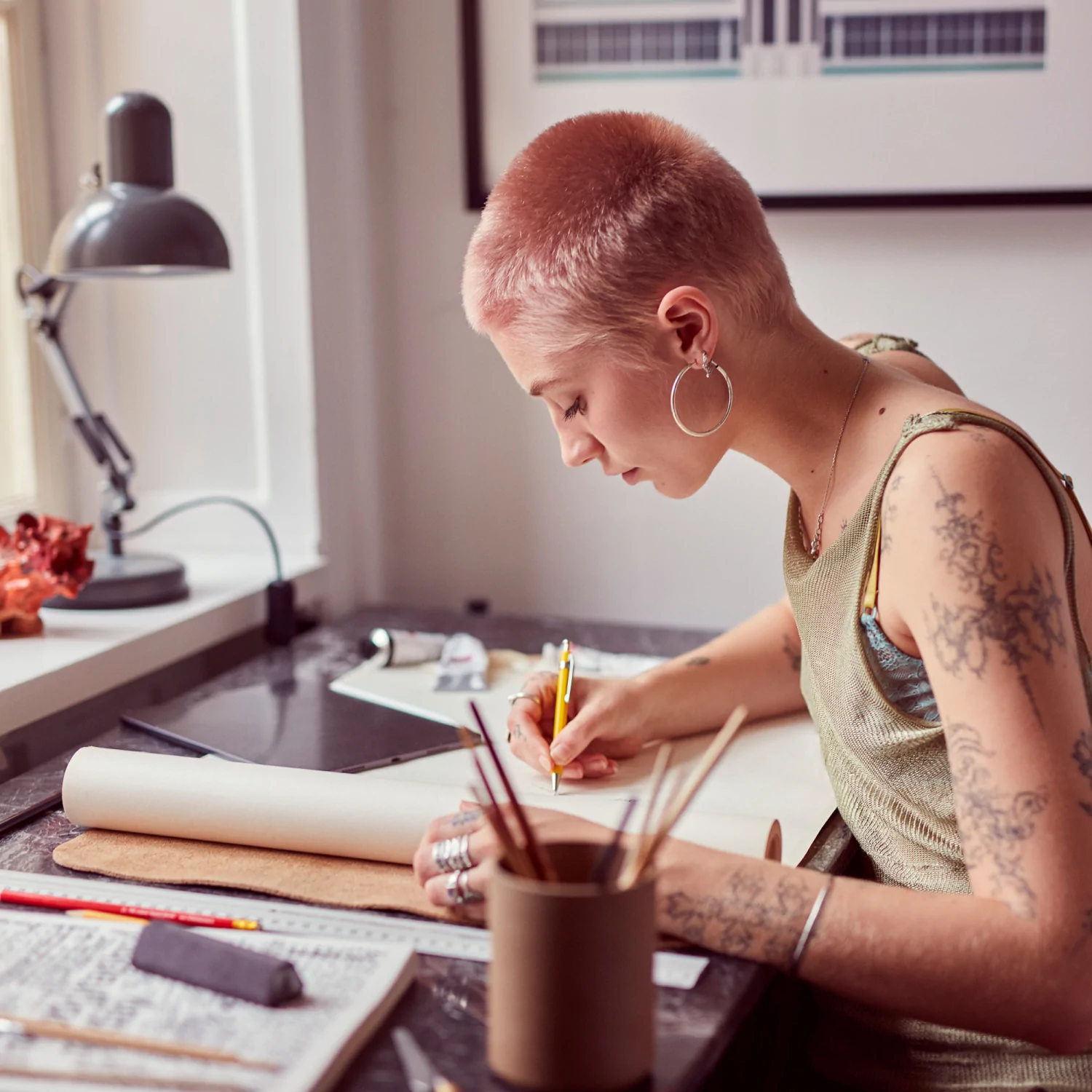 Frau mit pinken Haaren, die an einem Schreibtisch mit Kunstutensilien zeichnet, und eine Lampe.