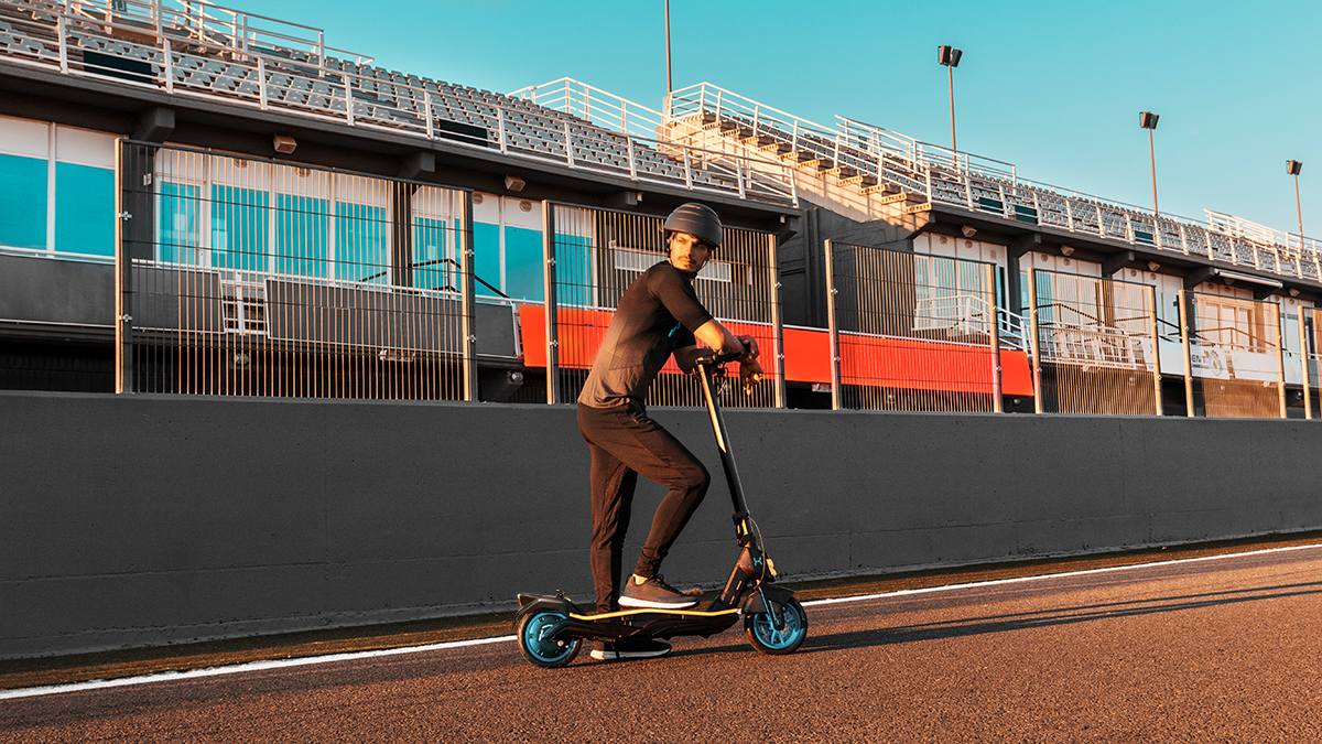 Un hombre monta un patinete eléctrico en una pista. Lleva un casco y ropa deportiva negra. El fondo muestra un estadio.