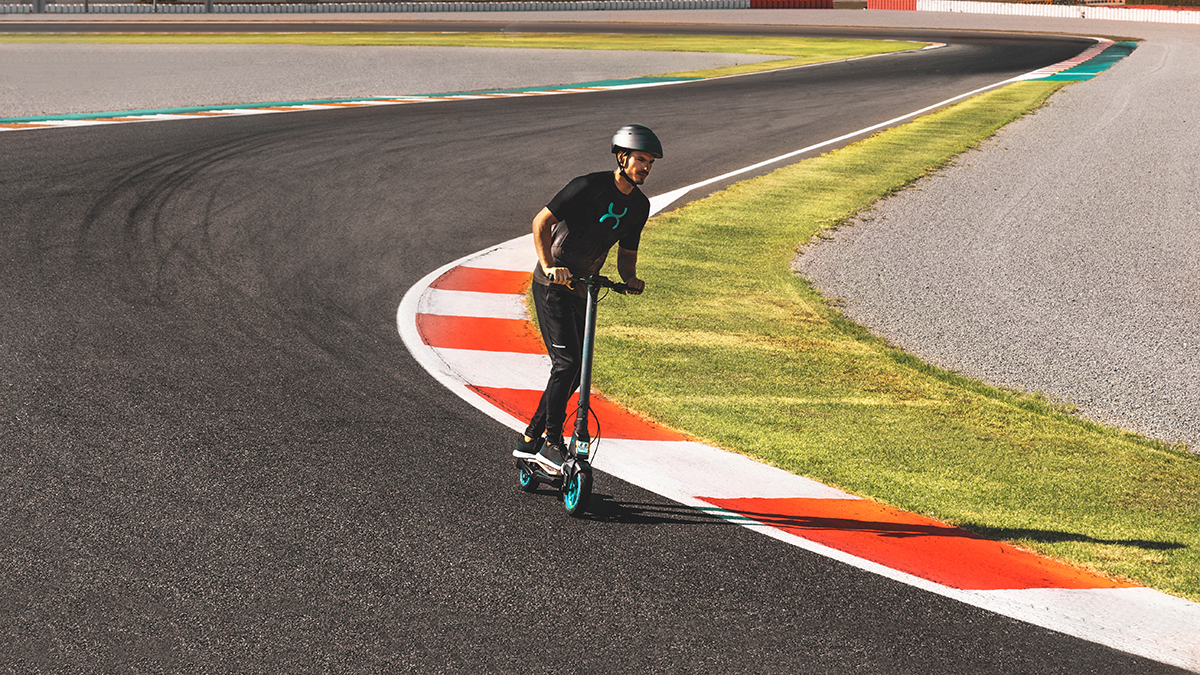 Un hombre monta un patinete eléctrico en una pista de carreras. Lleva casco y ropa negra. La pista tiene marcas rojas y blancas.