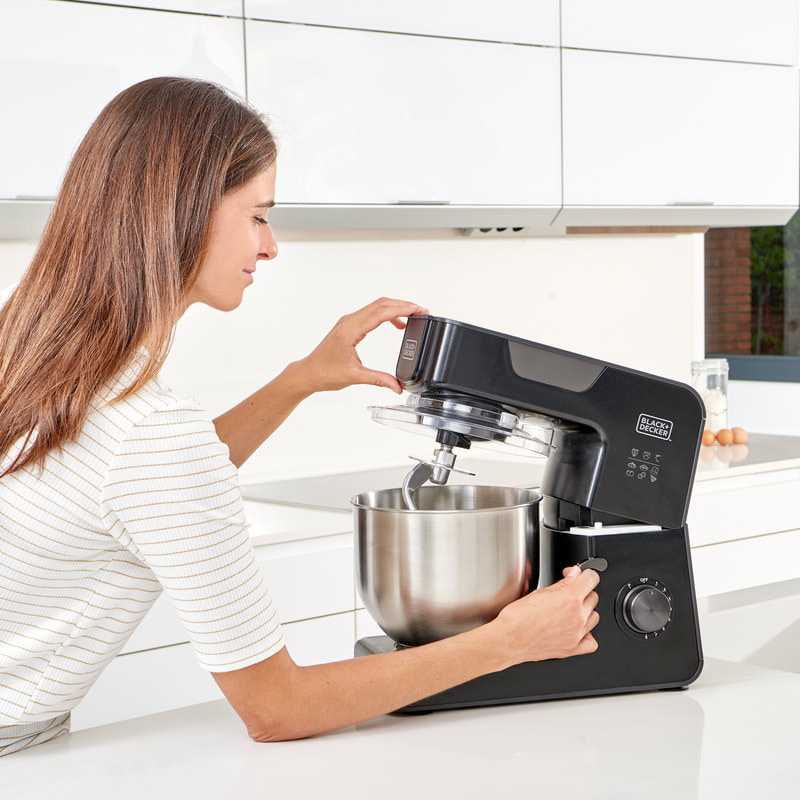 Nő fekete Black+Decker robotgépet használ fehér konyhában. Rozsdamentes acél tál belül.
