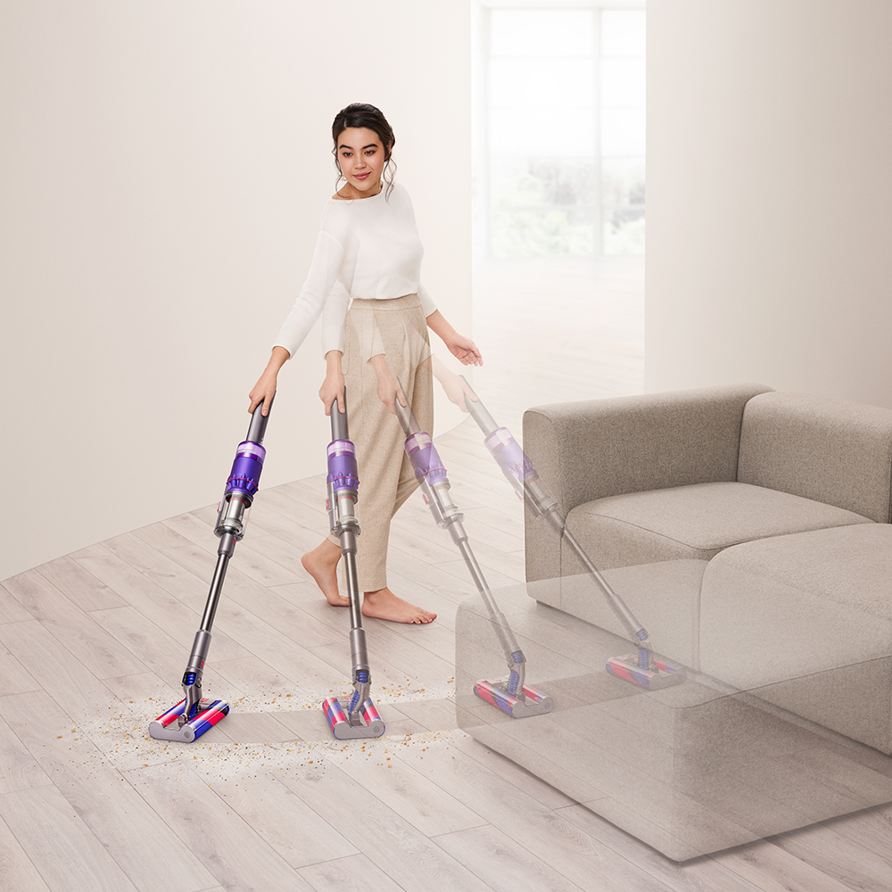 Vrouw die een stofzuiger gebruikt op een houten vloer. Een grijze bank op de achtergrond.