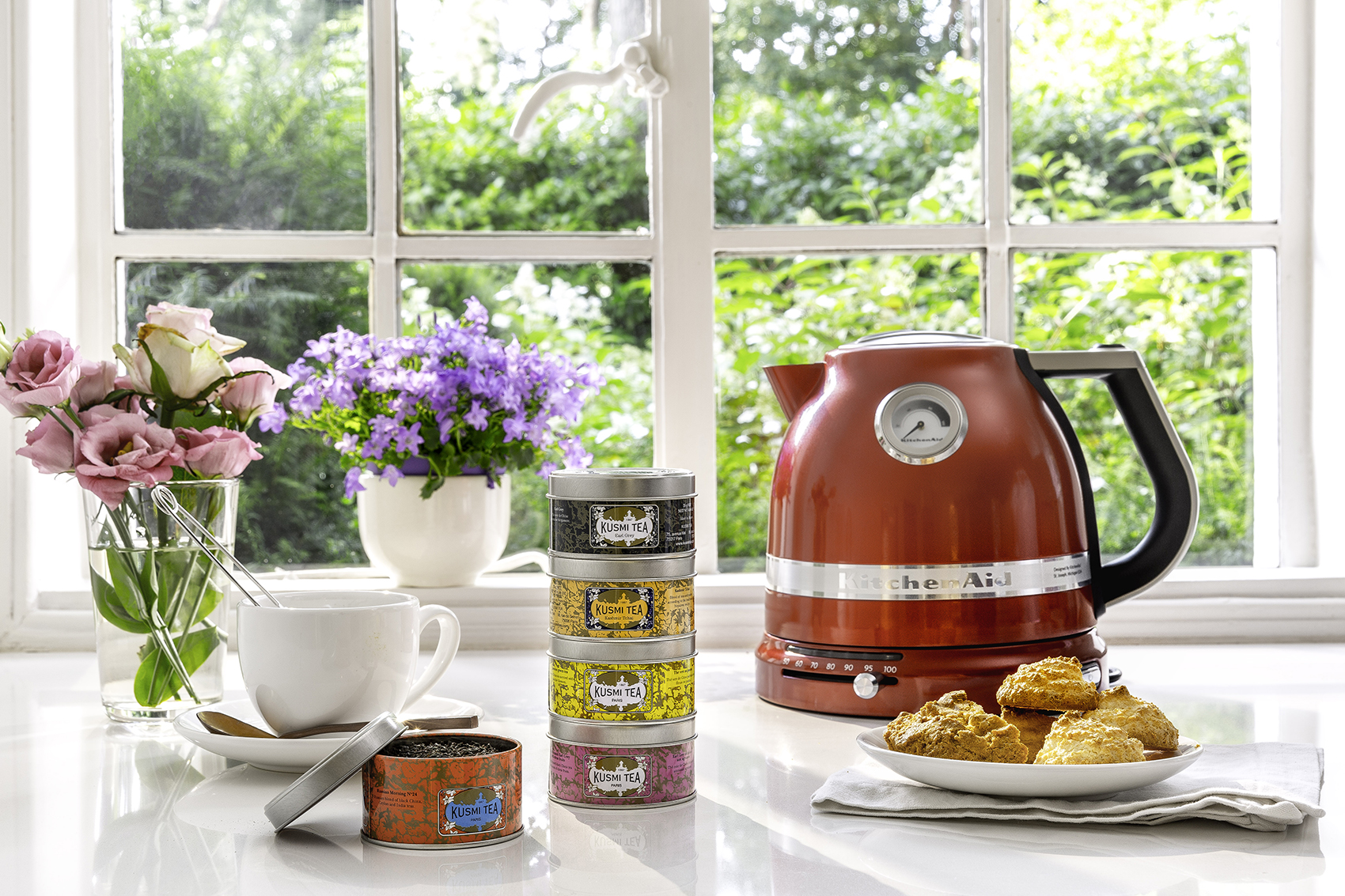 Una tetera KitchenAid roja con un dial, una taza blanca, latas de té y galletas en una encimera blanca cerca de una ventana.
