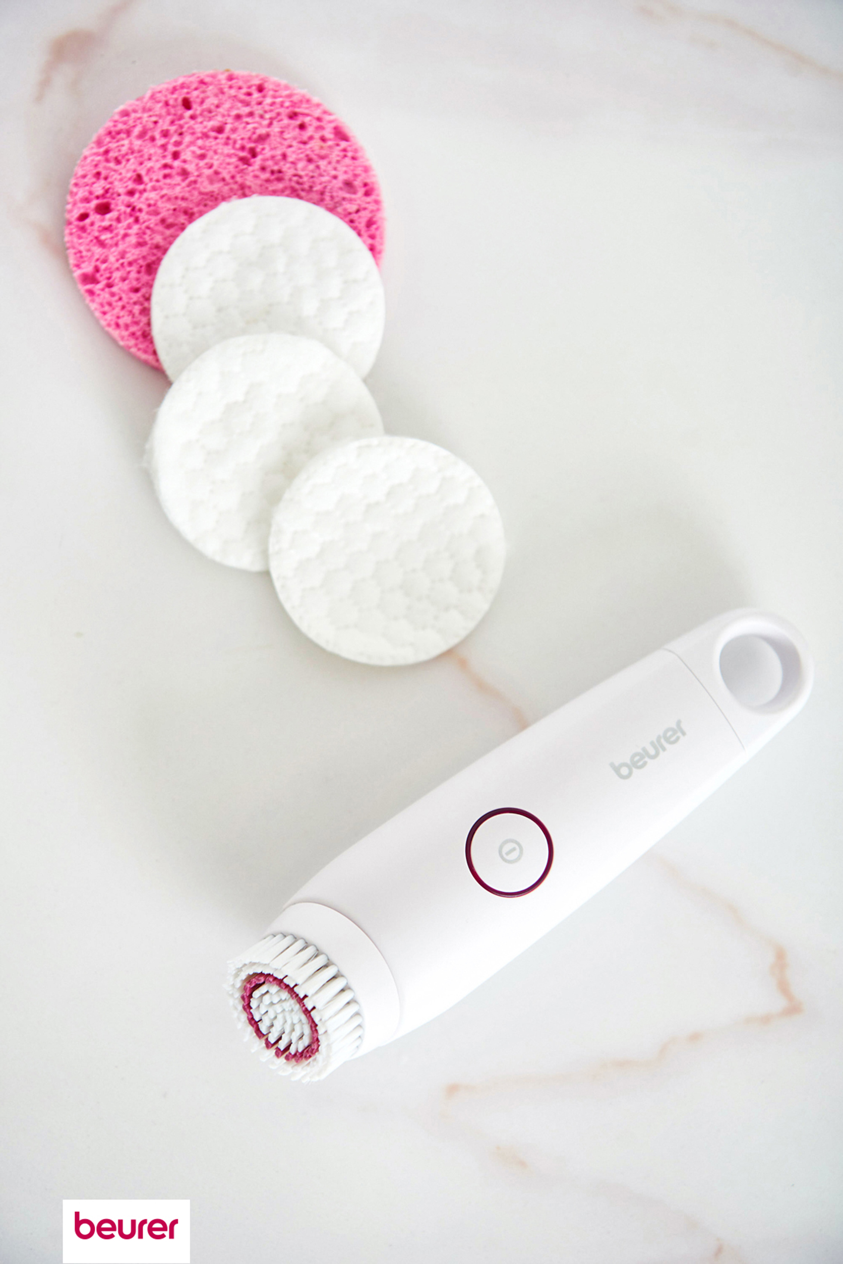 Un cepillo facial blanco con una esponja rosa y almohadillas de algodón blancas sobre mármol.