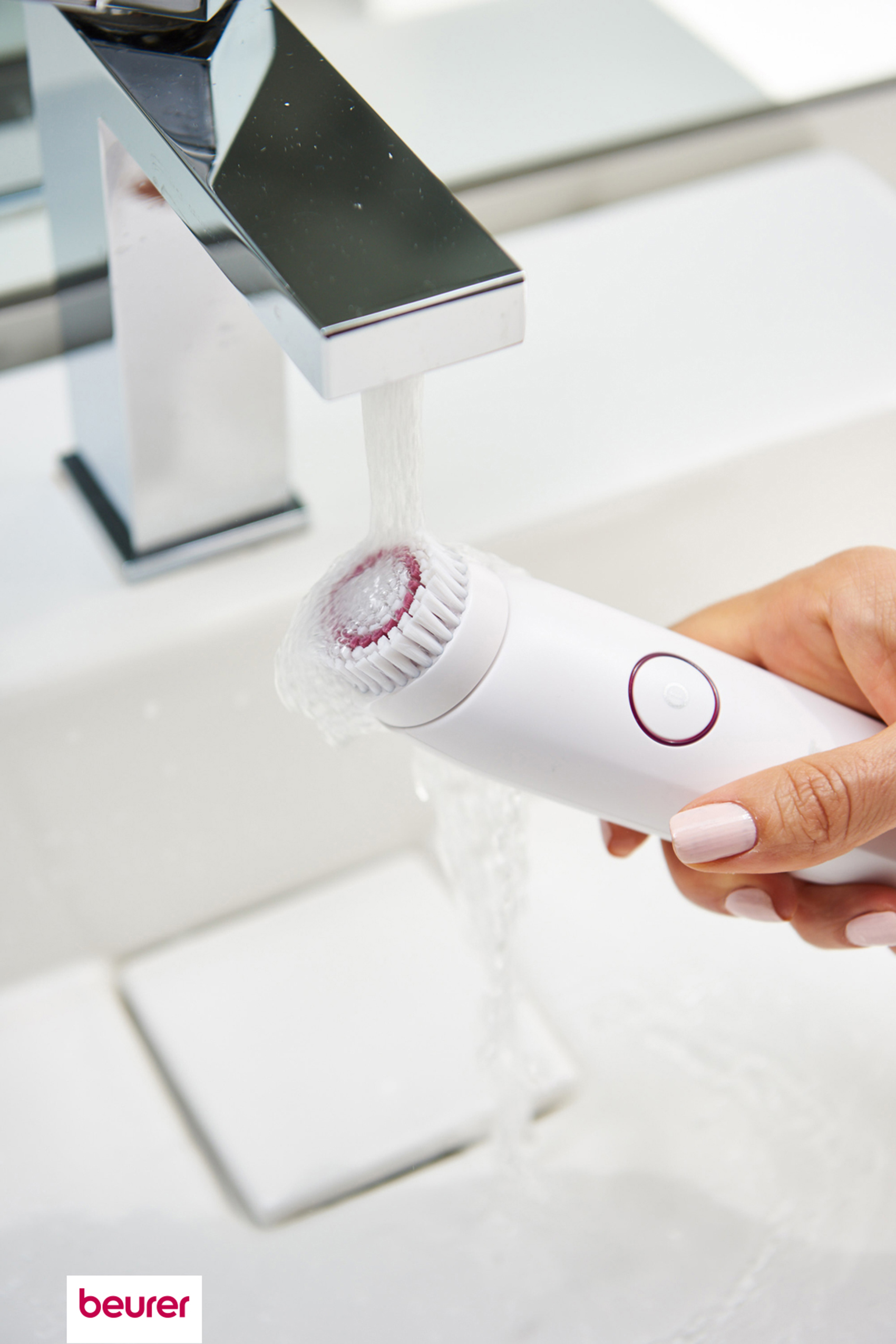 Una mano lava un cepillo facial blanco bajo agua corriente en un lavabo blanco.