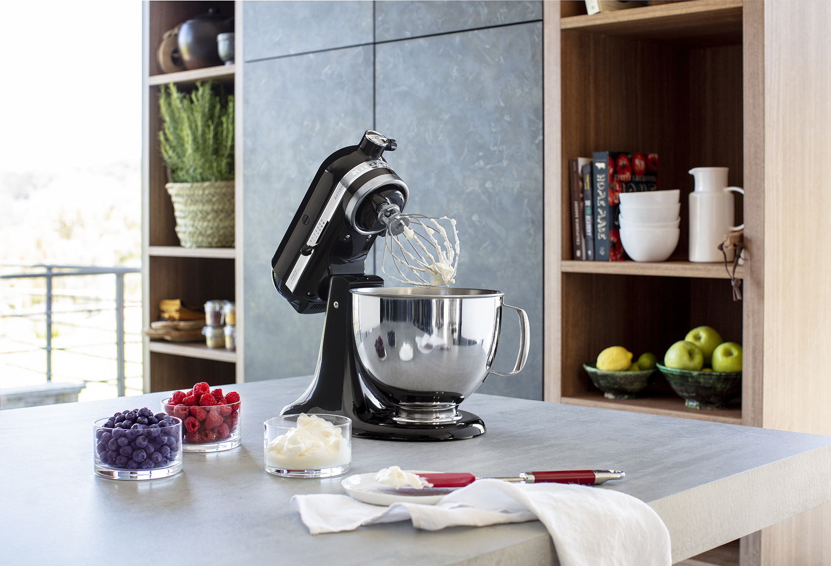 Batidora KitchenAid sobre encimera con ingredientes y ventana al fondo.