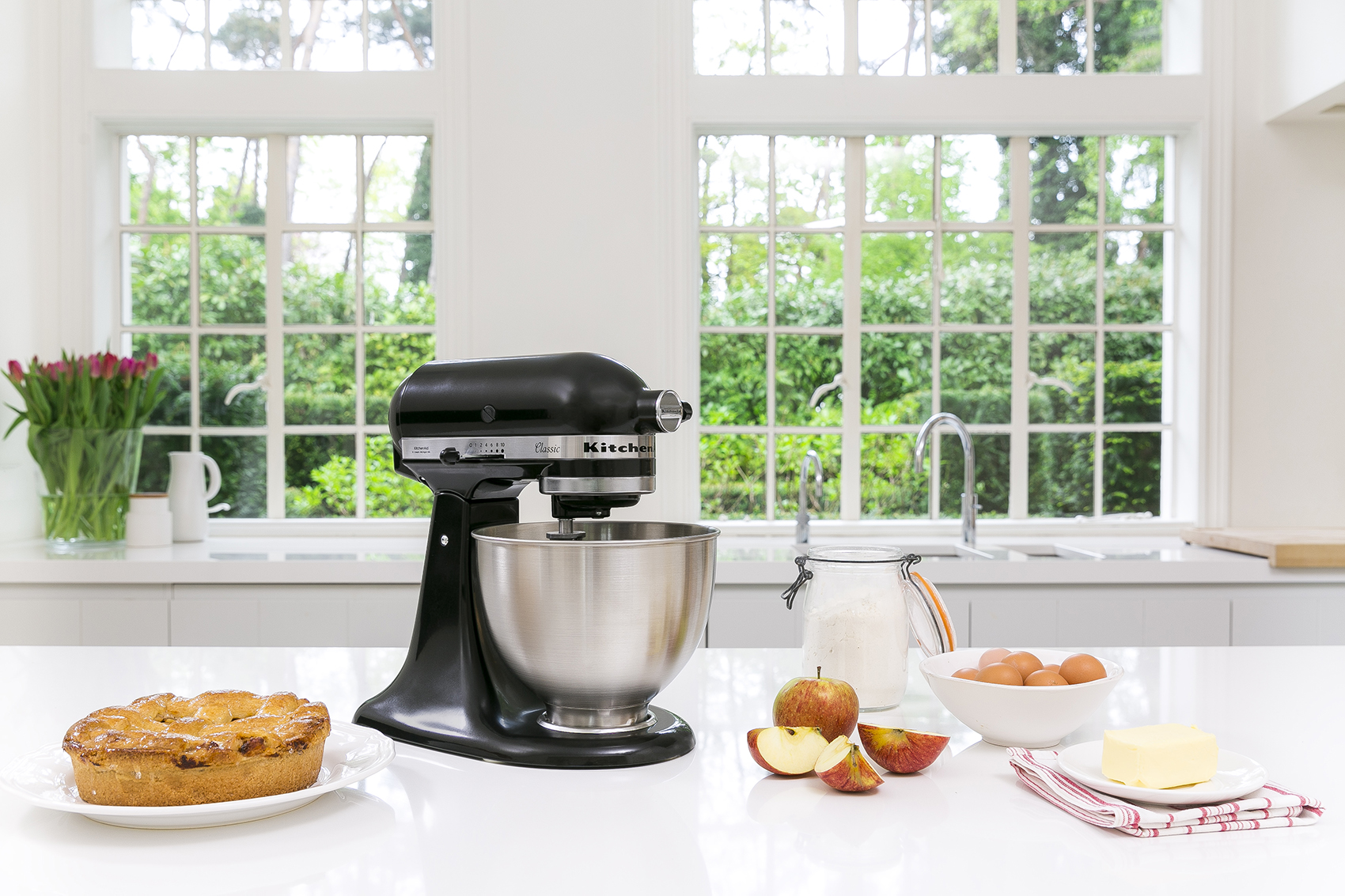 Batidora KitchenAid con pastel, manzanas e ingredientes sobre una encimera blanca cerca de ventanas.