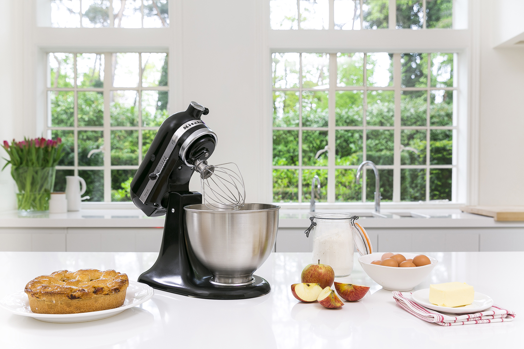 Una escena de cocina moderna con una batidora de pie negra, manzanas y otros ingredientes en una encimera blanca.