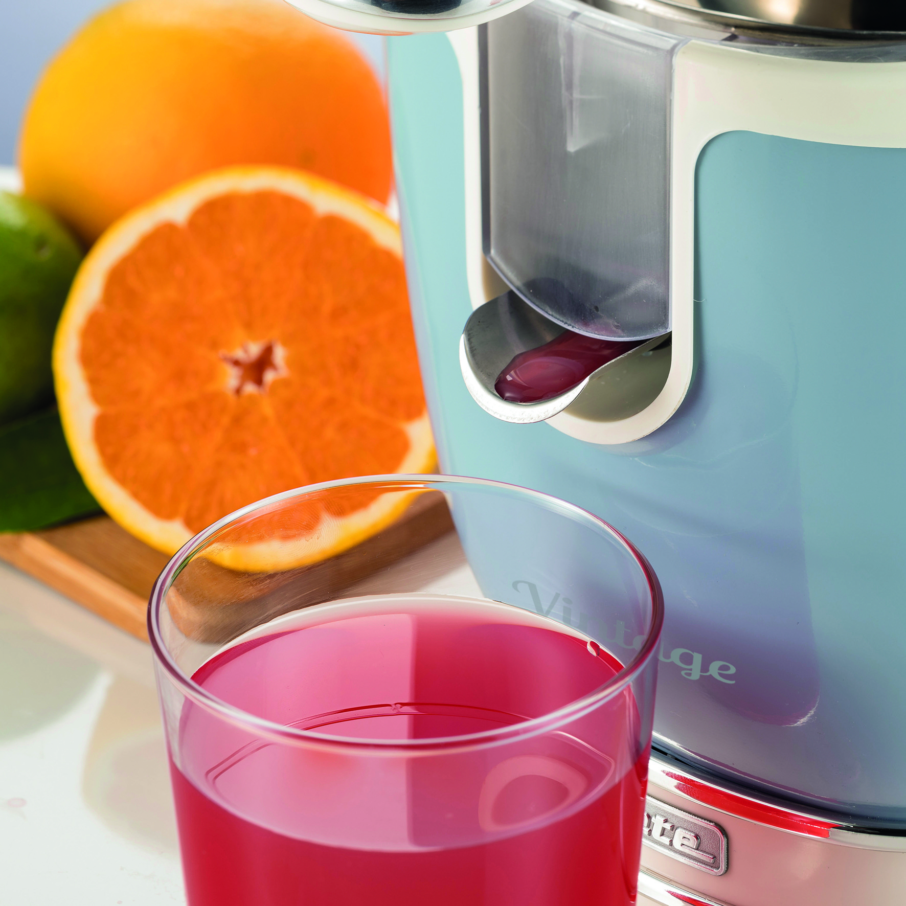 Una exprimidora, con un vaso de zumo y una naranja. Rodajas de naranja en el fondo.