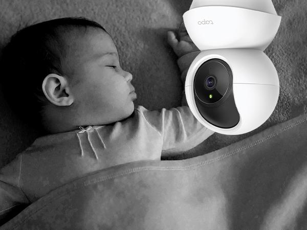 Ein Baby schläft unter einer Decke, eine weiße Überwachungskamera ist in der Nähe.