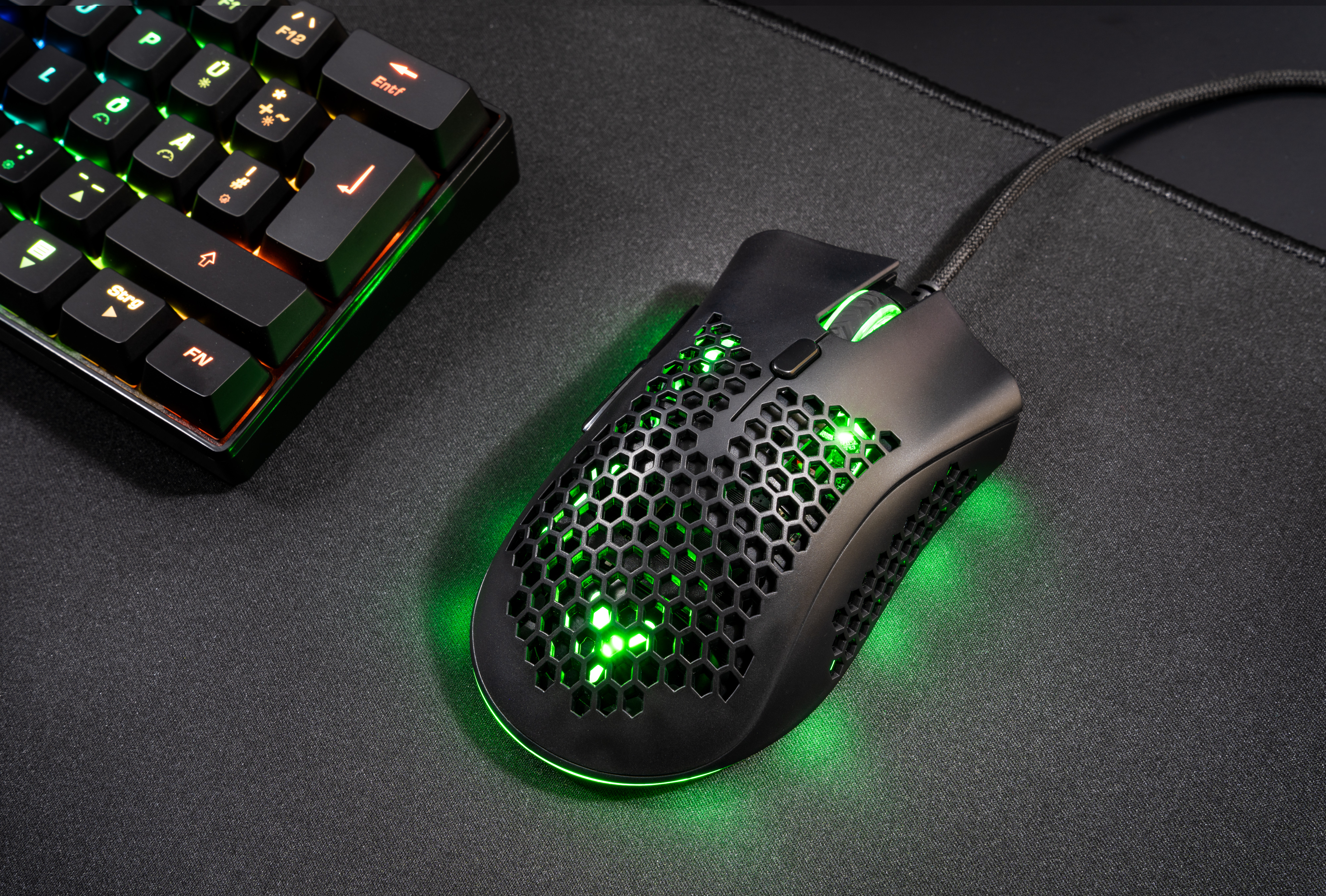 Eine schwarze Gaming-Maus mit grünen Lichtern auf einer schwarzen Matte. Im Hintergrund befindet sich eine Tastatur mit bunten Lichtern.