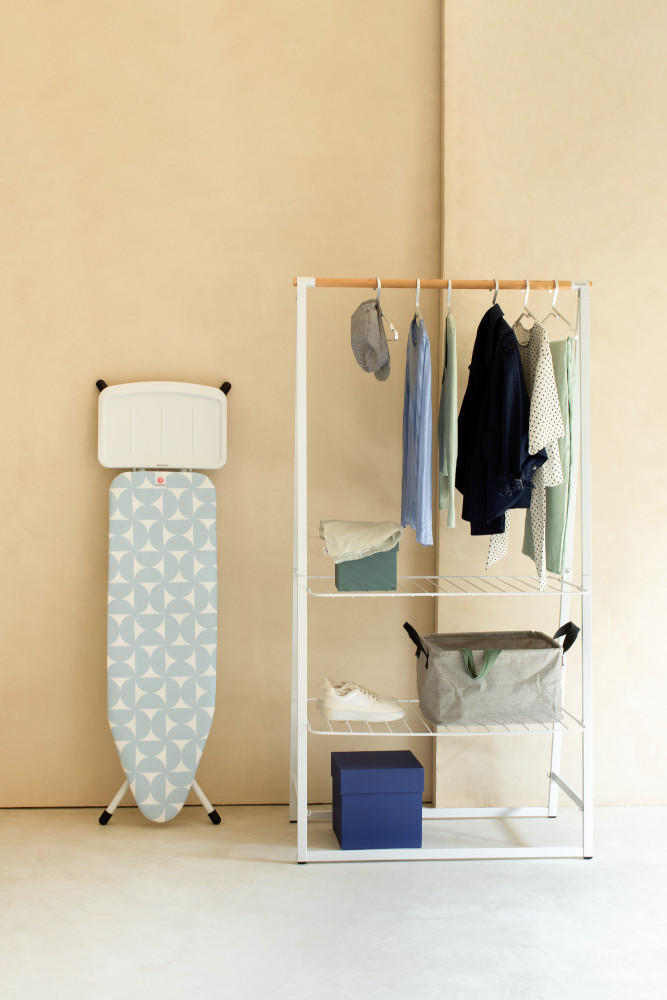 Een witte kledingrek met hangende kleding, een strijkplank en een blauwe opbergdoos.