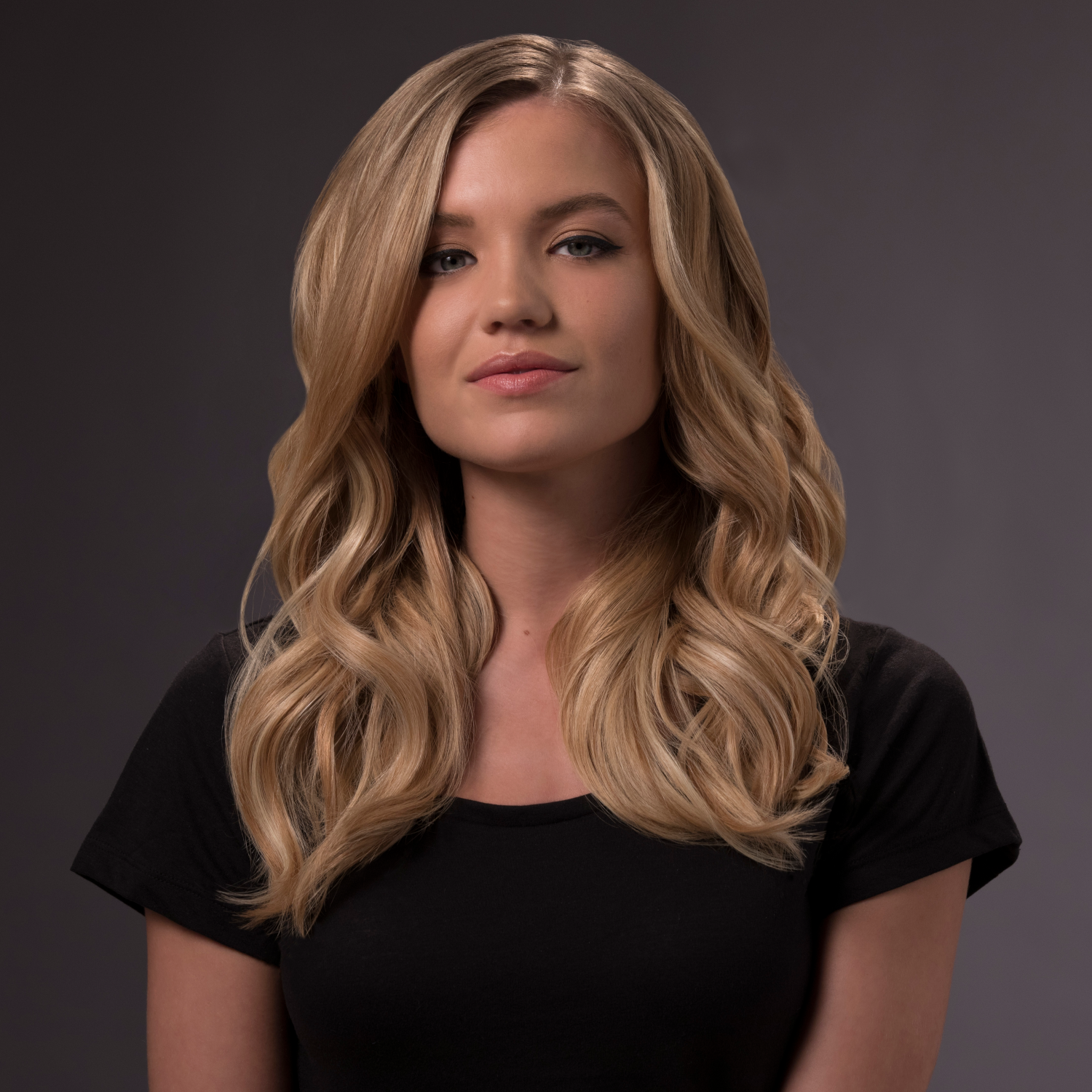Blonde vrouw in een zwart shirt. Lang, golvend haar. Neutrale grijze achtergrond.