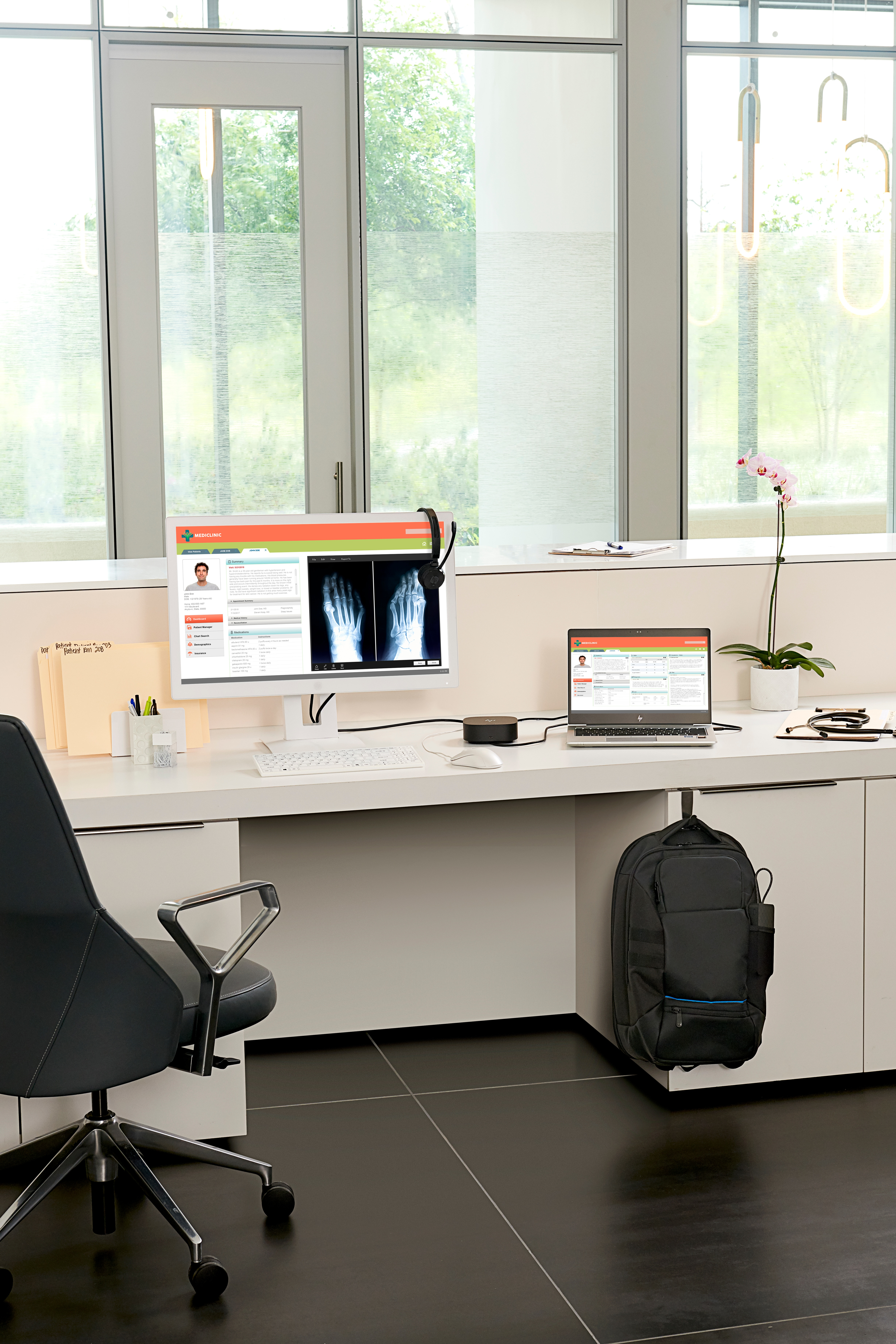 Bürotisch mit Computer, Röntgenanzeige, Laptop und Zubehör. Fenster und Tür im Hintergrund.
