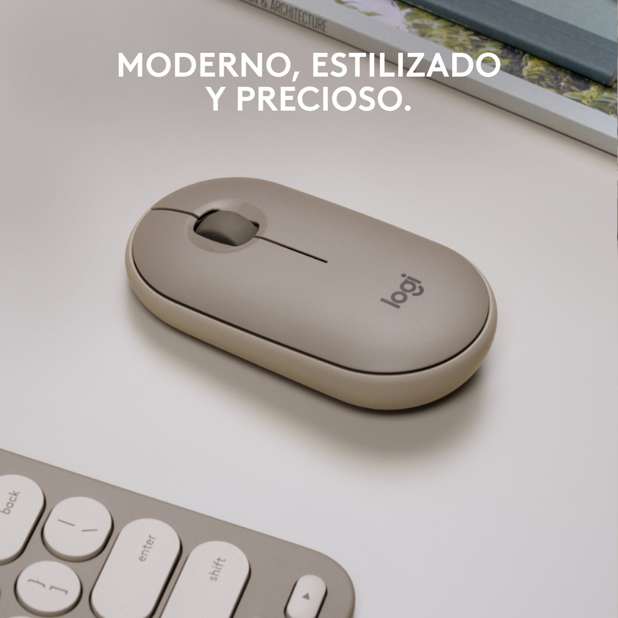 Un ratón Logitech gris claro sobre un escritorio blanco con un teclado en primer plano y un libro en el fondo.