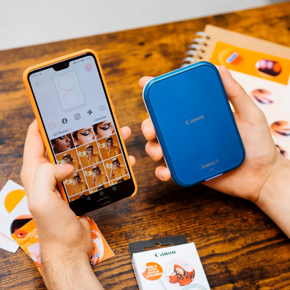 Een persoon houdt een telefoon en een blauwe Canon Zoemini 2 printer met stickers vast.