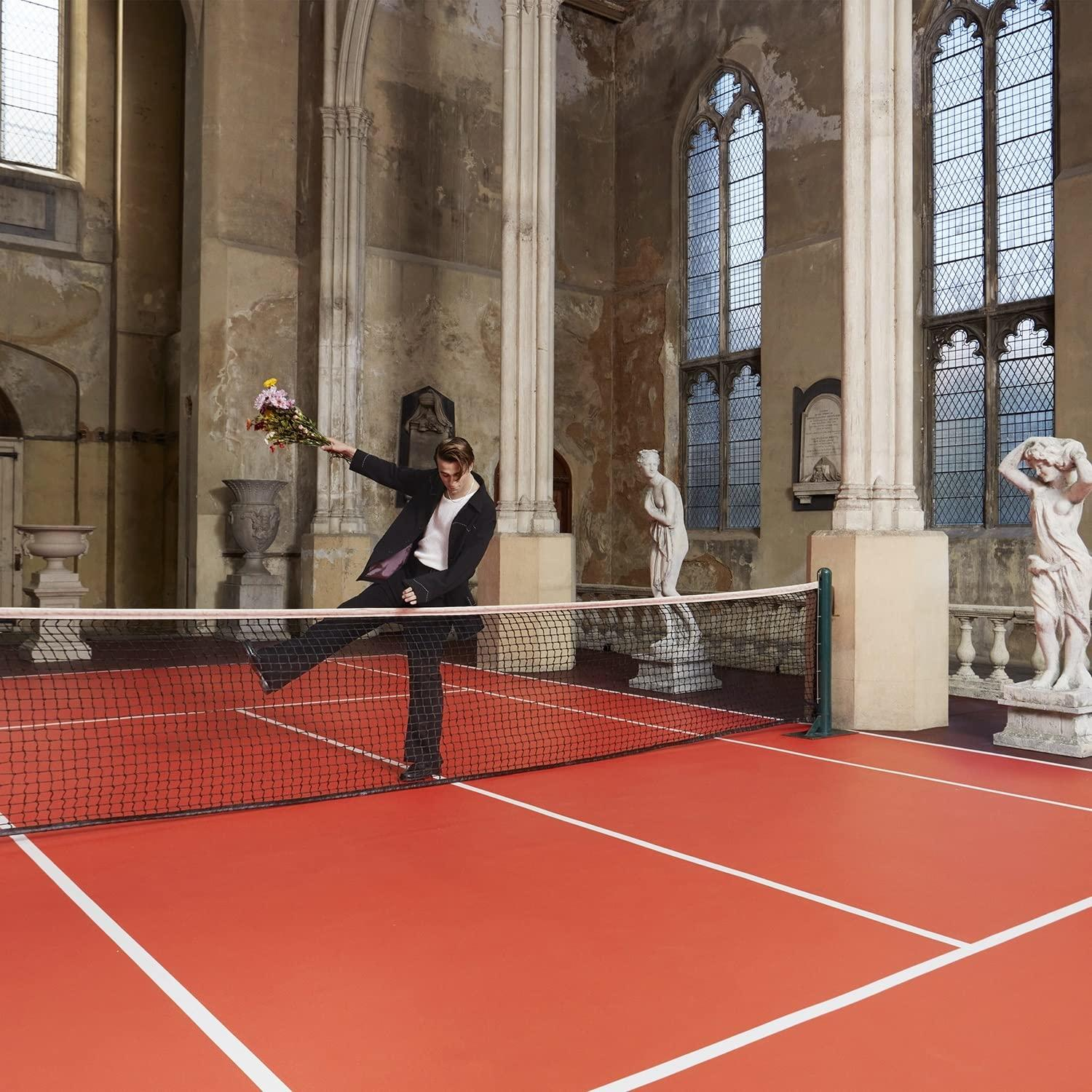 Mann im Anzug tritt Blumenball auf rotem Tennisplatz. Statuen und Fenster im Hintergrund.