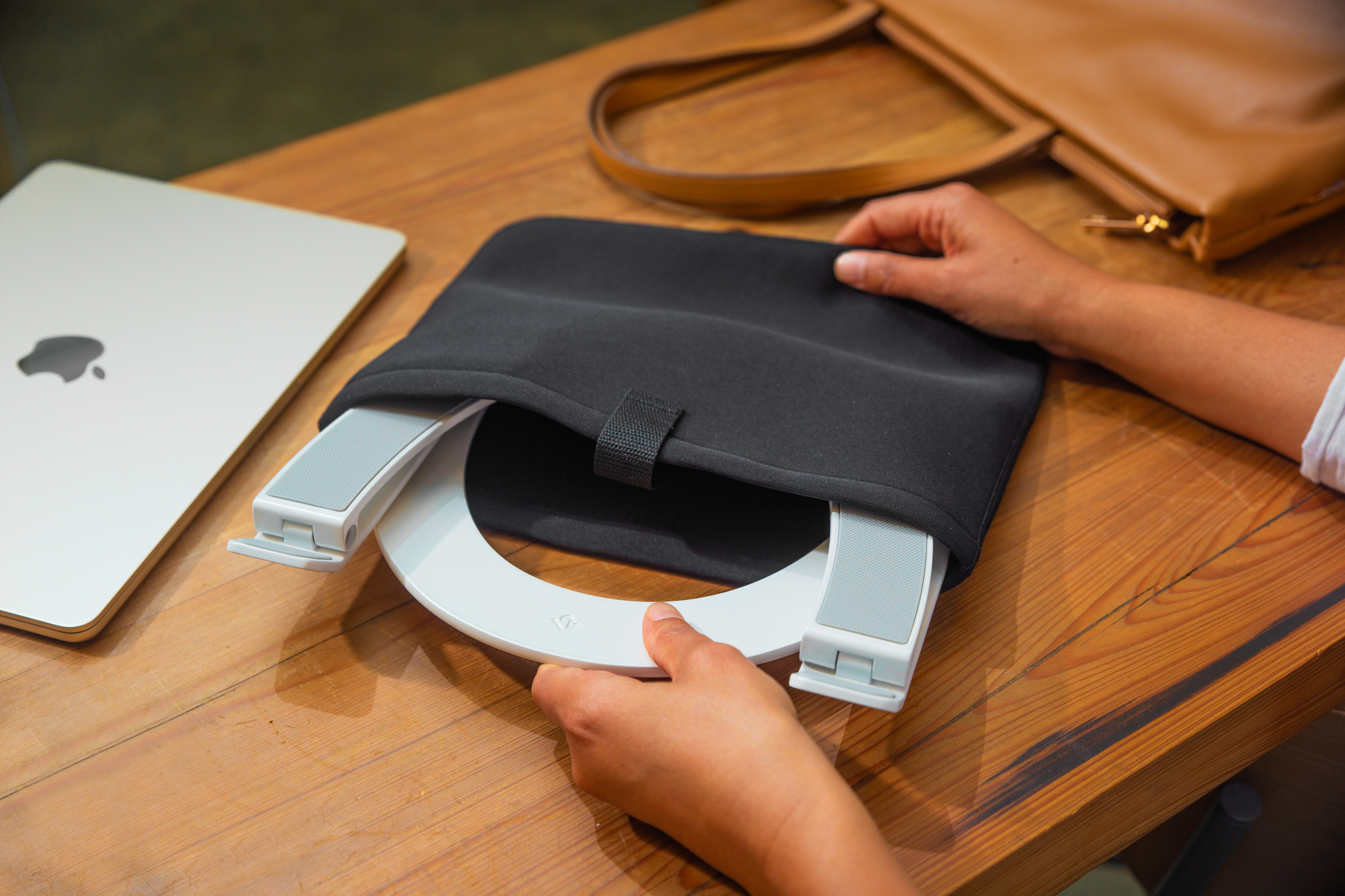 Hände halten einen schwarzen Laptopständer auf einem Holztisch, mit Laptop und Handtasche im Hintergrund.