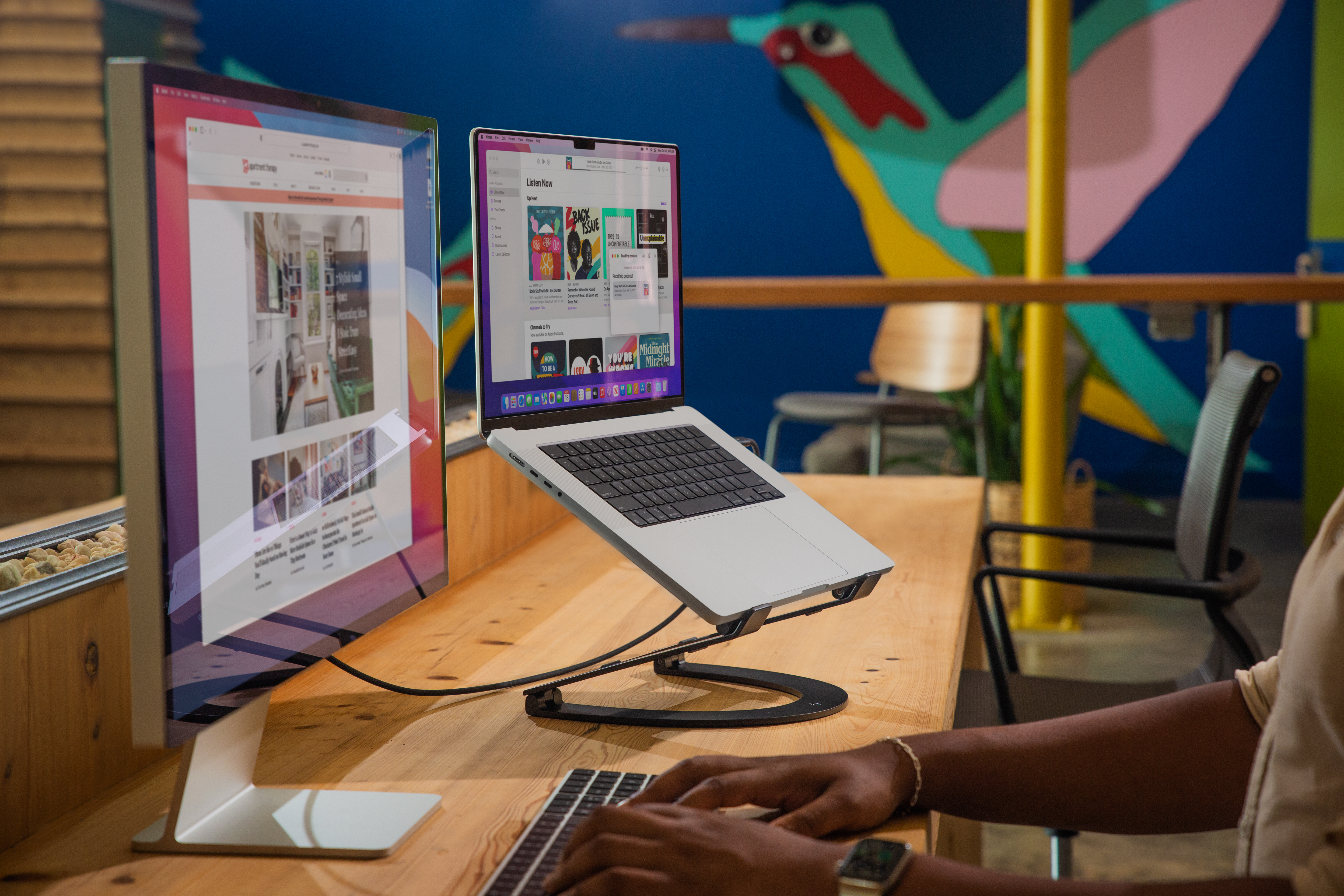 Eine Person tippt auf einer Tastatur. Zwei Bildschirme zeigen Websites. Ein Laptop steht auf einem Ständer. Hintergrund: bunte Wandkunst.
