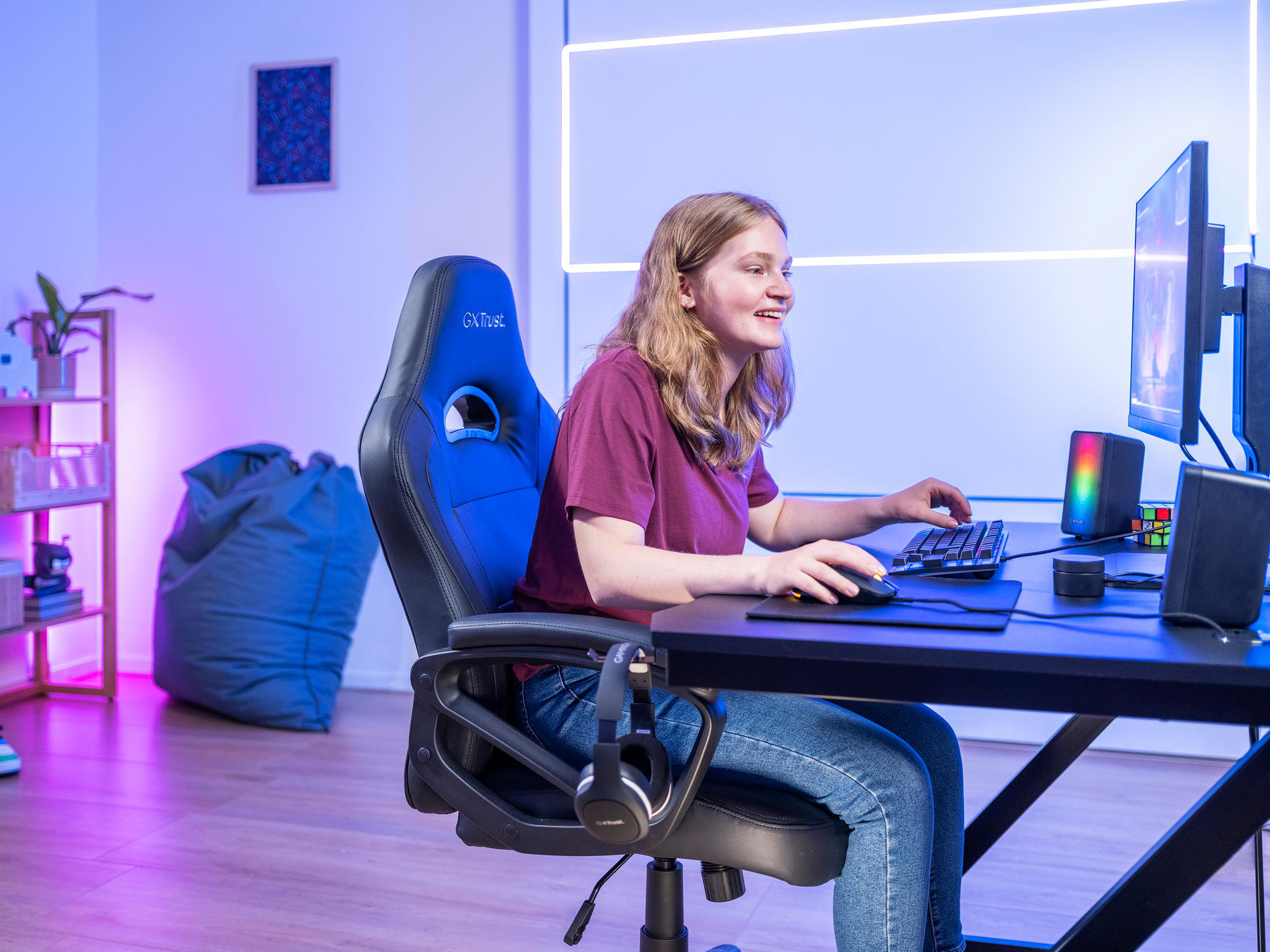 Eine junge Frau sitzt in einem Gaming-Stuhl und lächelt, während sie an einem beleuchteten Schreibtisch am Computer spielt.