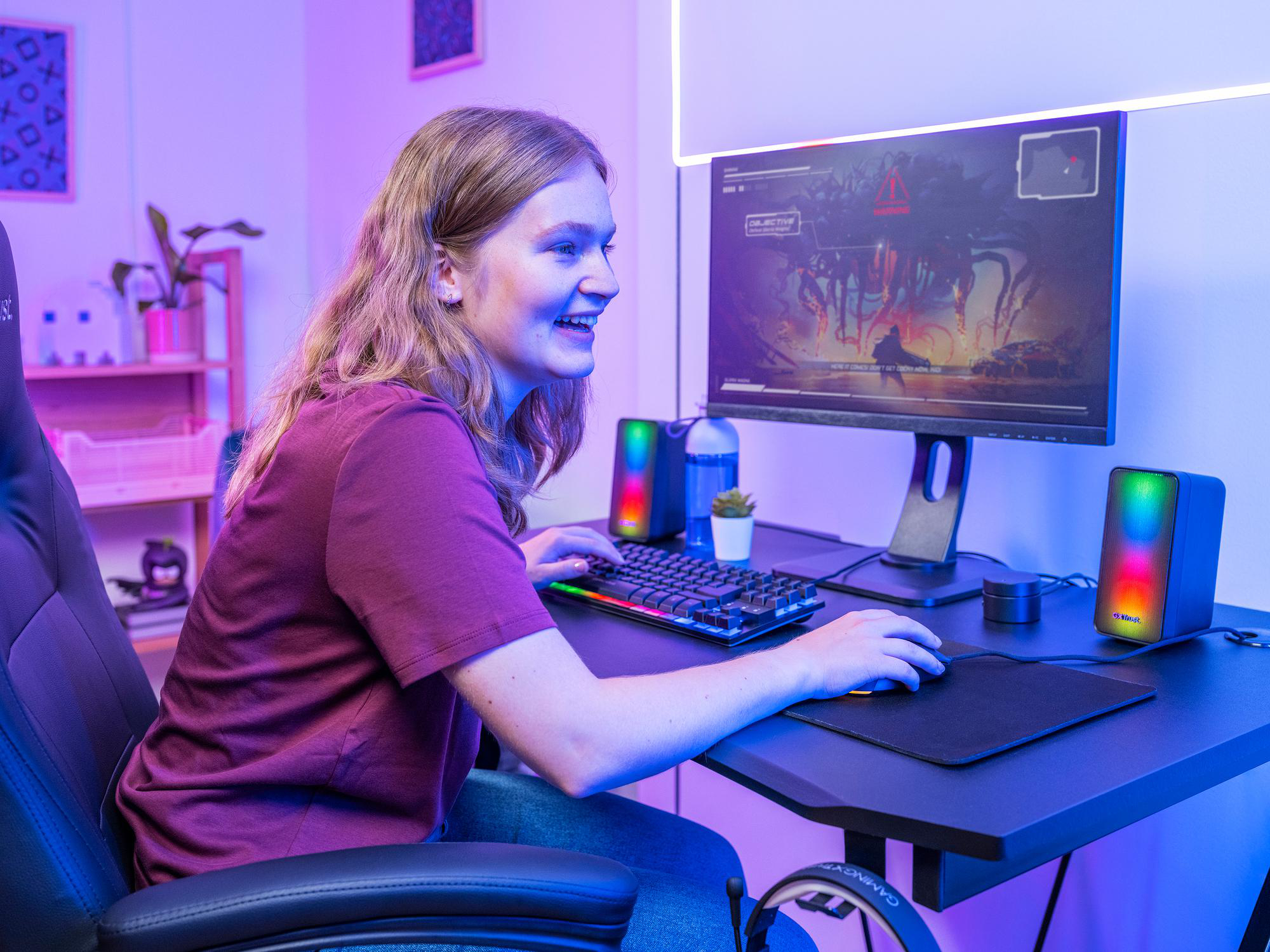 Eine junge Frau sitzt vor einem beleuchteten PC-Bildschirm und spielt ein Spiel in einem lila beleuchteten Raum.