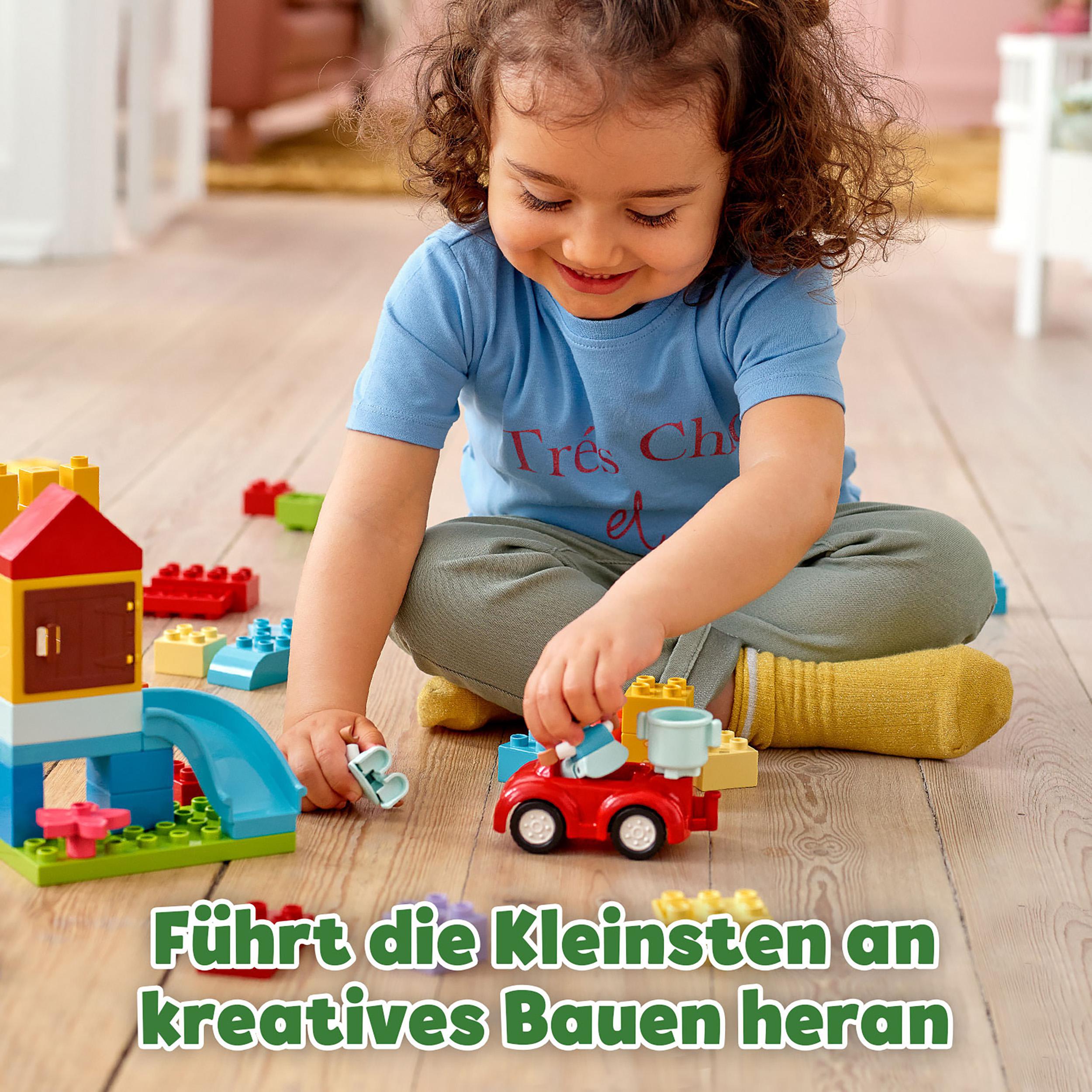 Ein Kind spielt mit Lego-Steinen. Ein rotes Auto und ein Gebäude stehen auf einem Holzboden.