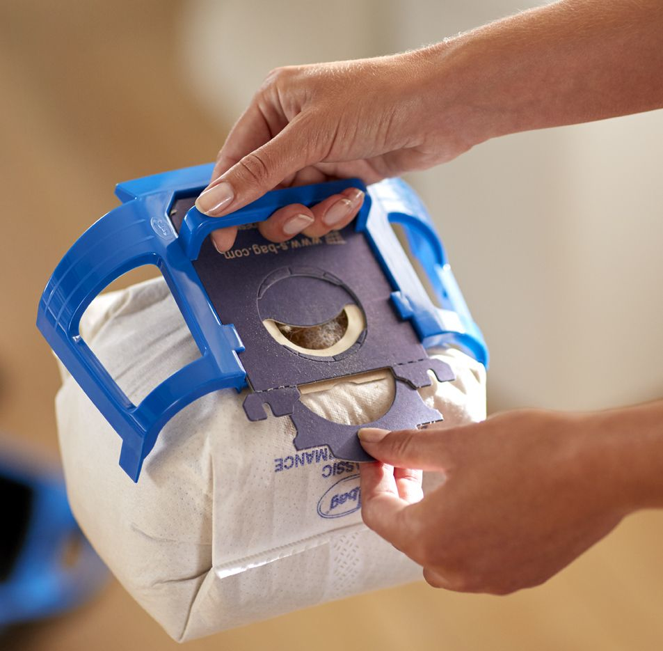 Hände halten einen blauen Staubsaugerbeutelhalter und setzen einen neuen Staubsaugerbeutel ein. Die Tasche ist beige, mit weißem Text.