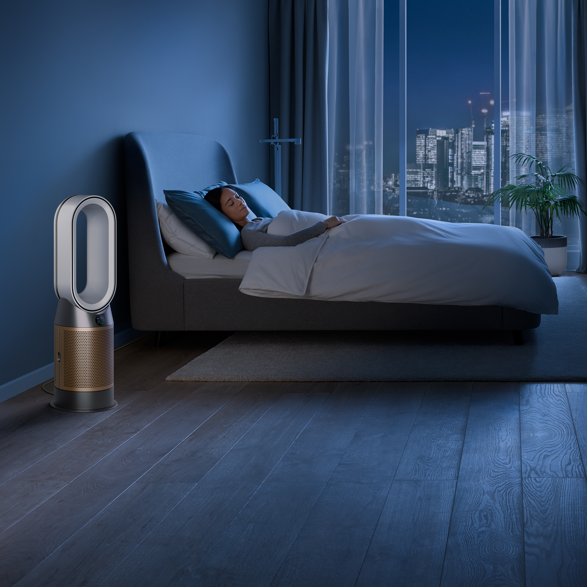 Une personne dort dans le lit. Un purificateur d'air est à gauche. Lumières de la ville à l'extérieur.