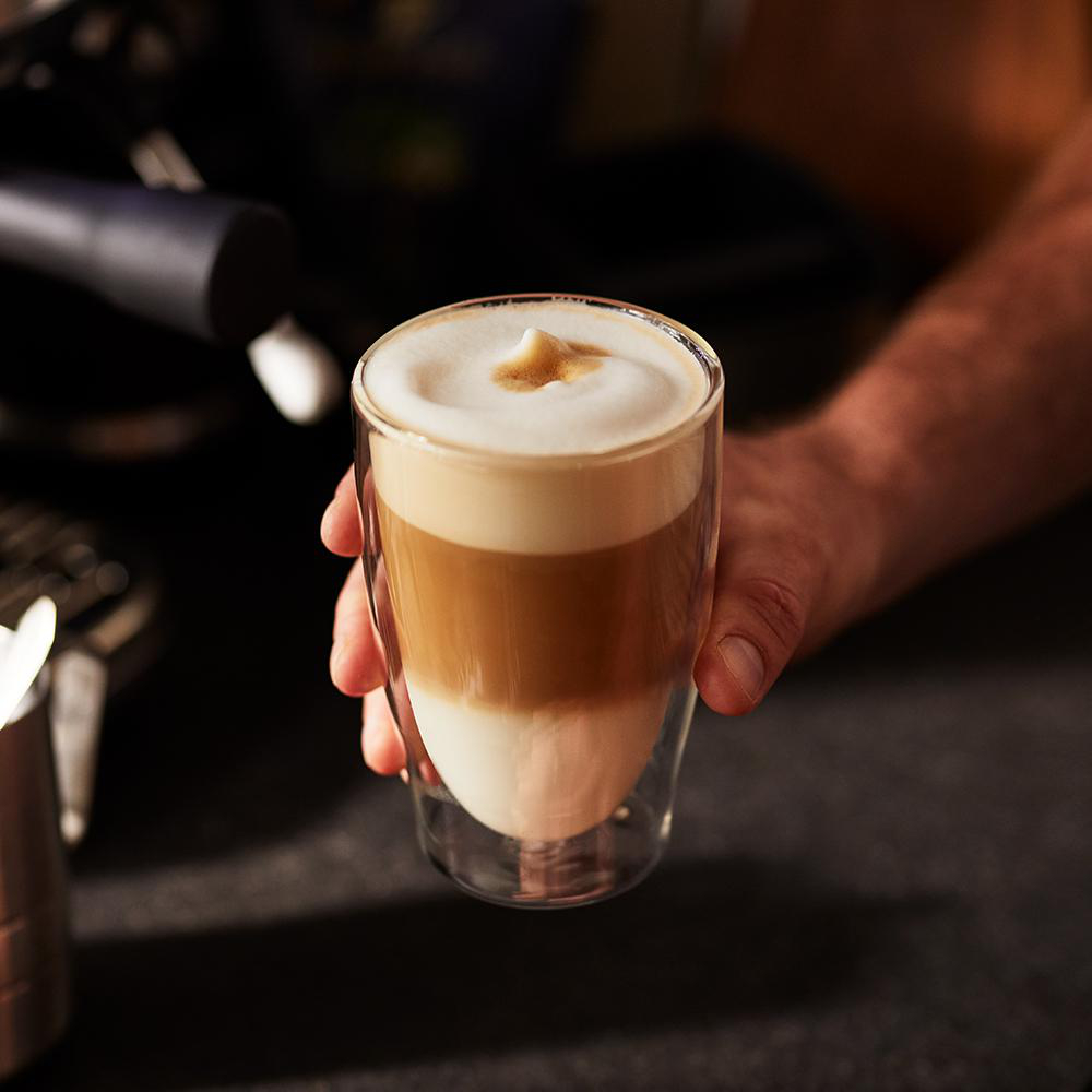 Eine Hand hält ein Glas mit geschichtetem Kaffeegetränk: weißer Schaum, hellbraun und weiß.