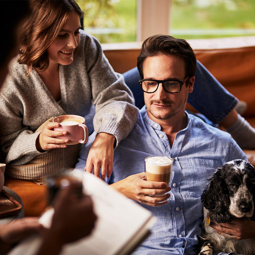 Ein Paar trinkt Kaffee, ein Hund sitzt neben dem Mann. Eine weitere Person liest ein Buch.