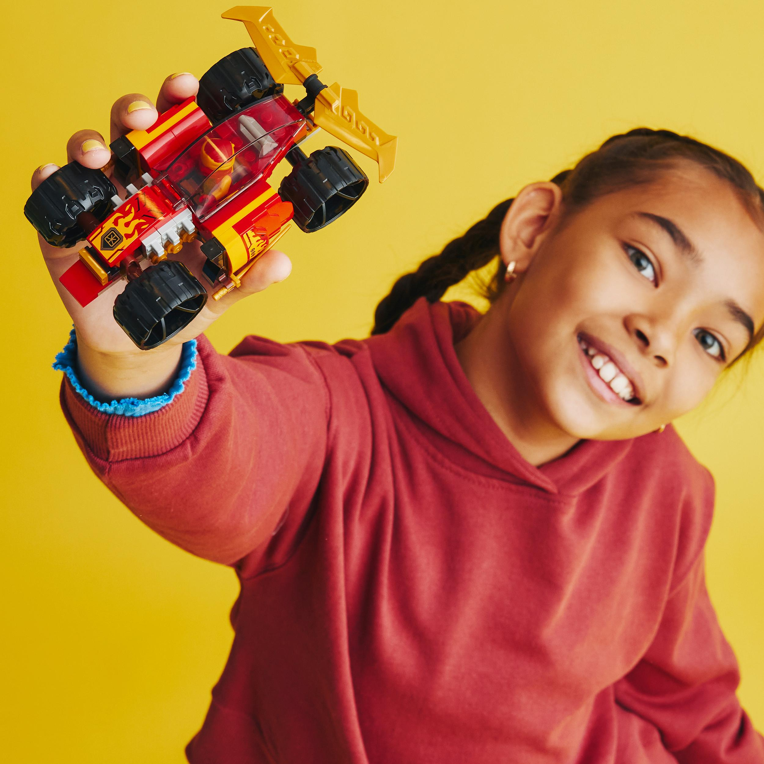 Ein Mädchen hält ein rot-gelbes Lego-Rennauto und lächelt. Der Hintergrund ist gelb.