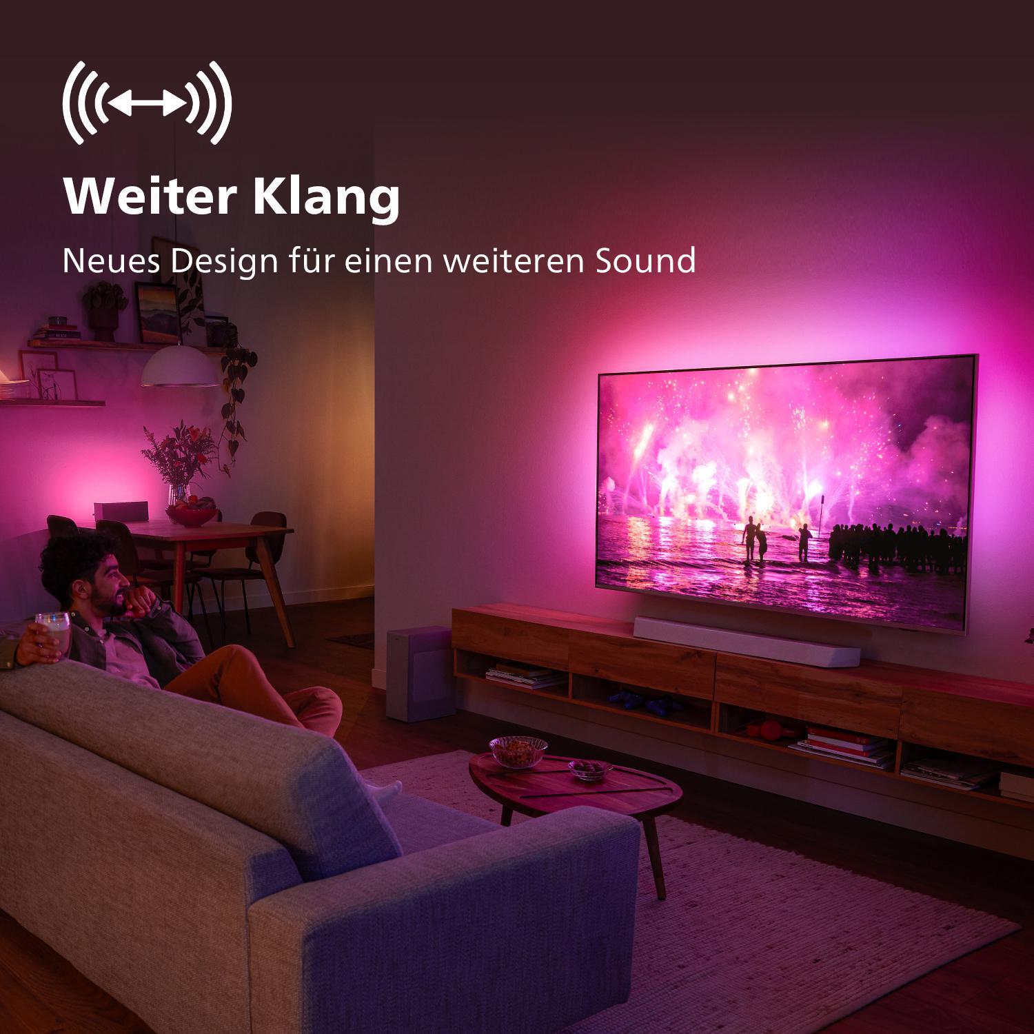 Ein Wohnzimmer mit einem Fernseher, der farbenfrohe Lichter anzeigt. Eine Person sitzt mit einem Getränk auf dem Sofa.