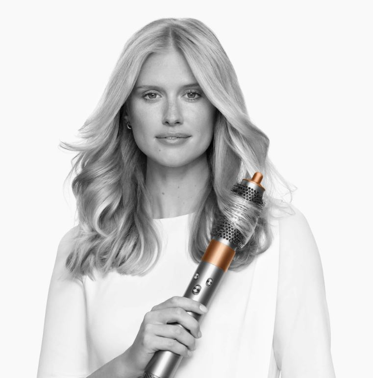 Donna che tiene un'arricciacapelli Dyson. Ha lunghi capelli biondi e sorride.