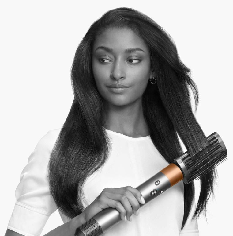 Una donna con lunghi capelli scuri tiene in mano uno strumento per lo styling dei capelli Dyson, lo sfondo è bianco.