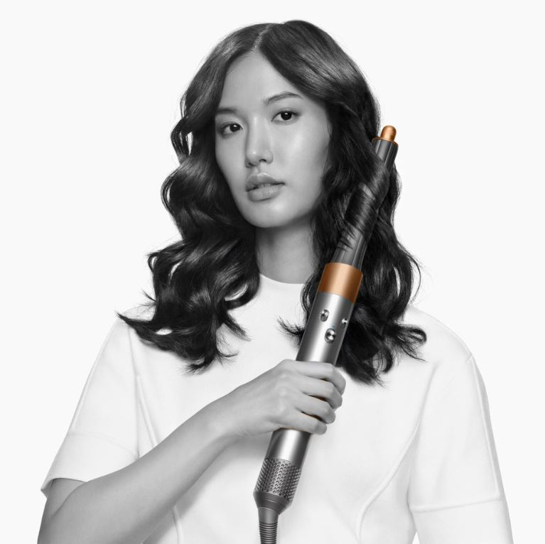 Una donna con i capelli scuri li arriccia con uno styler per capelli argentato e ramato. Indossa una camicia bianca.