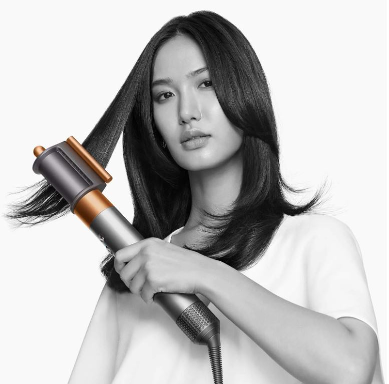 Donna usa uno strumento per lo styling dei capelli sui suoi capelli scuri su sfondo bianco. Lo strumento è grigio e bronzo.