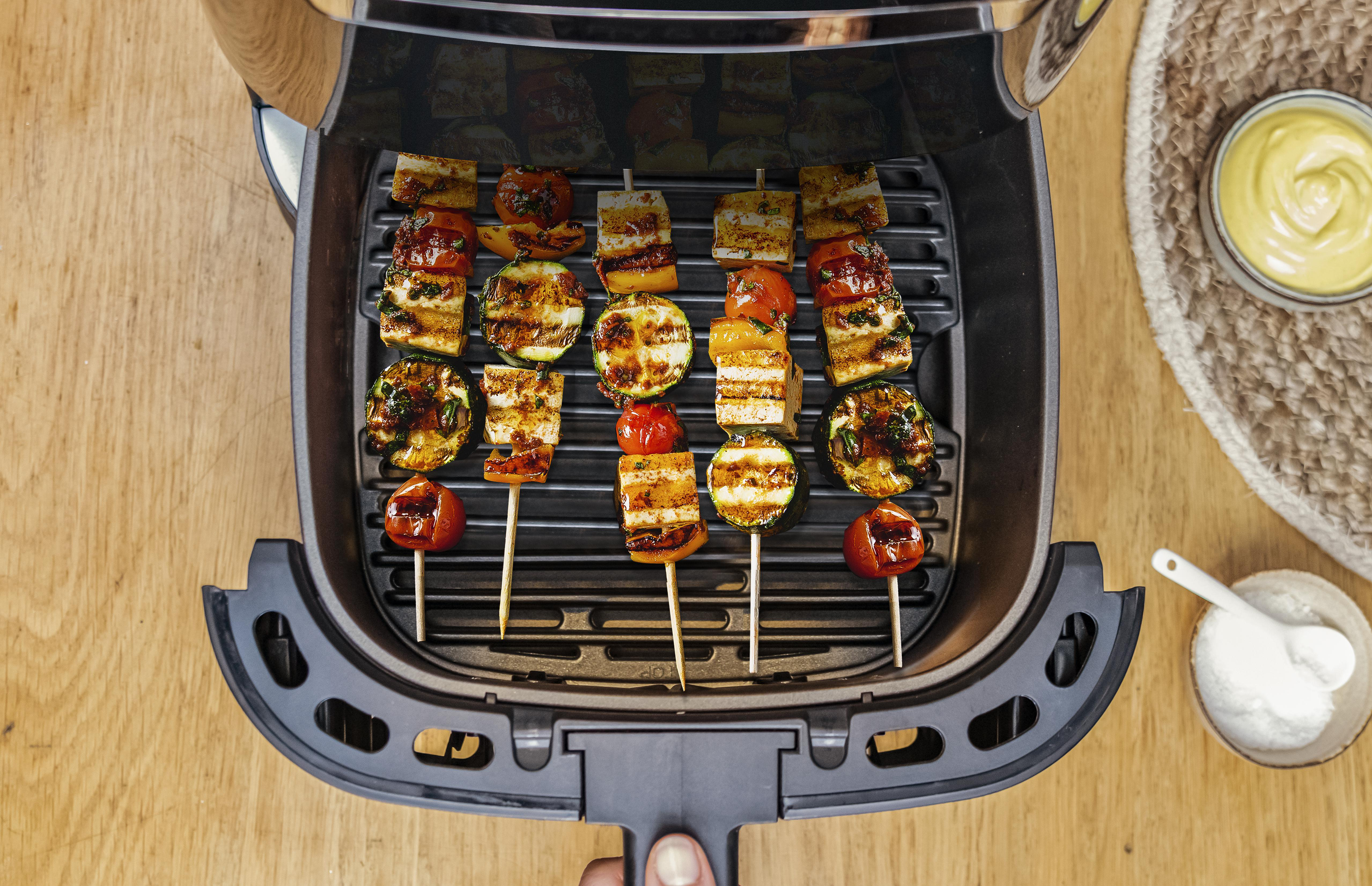 Eine Heißluftfritteuse mit Spießen mit gegrilltem Gemüse und einer Schüssel Soße auf einem Holztisch.