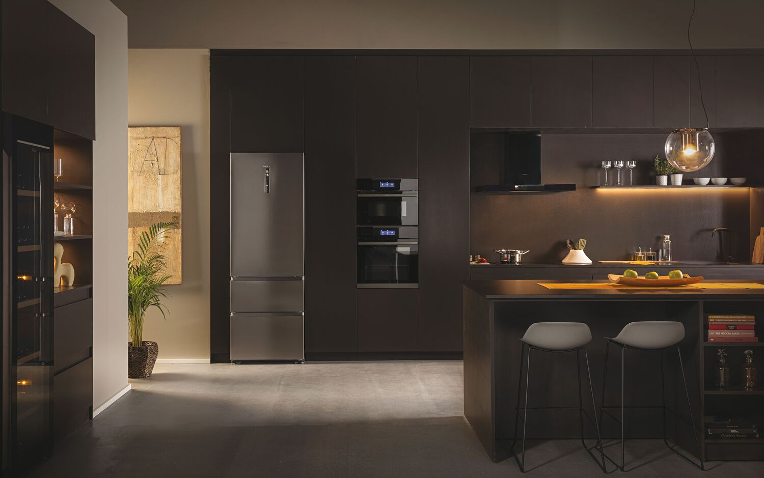 Interior de cocina moderna. Refrigerador, hornos, mostrador y taburetes. Colores neutros e iluminación cálida.