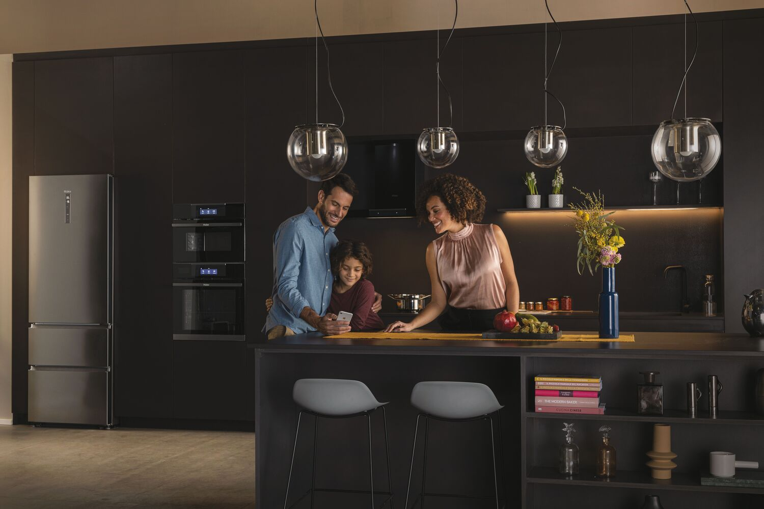 Una familia en una cocina, mirando un teléfono. Se ven un refrigerador y electrodomésticos.