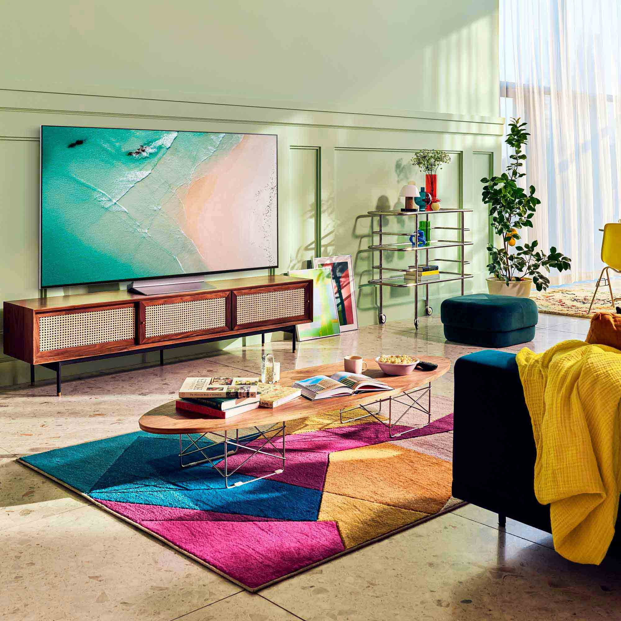 Una sala de estar con un televisor, un sofá y una alfombra colorida. Hay una planta y una mesa auxiliar.