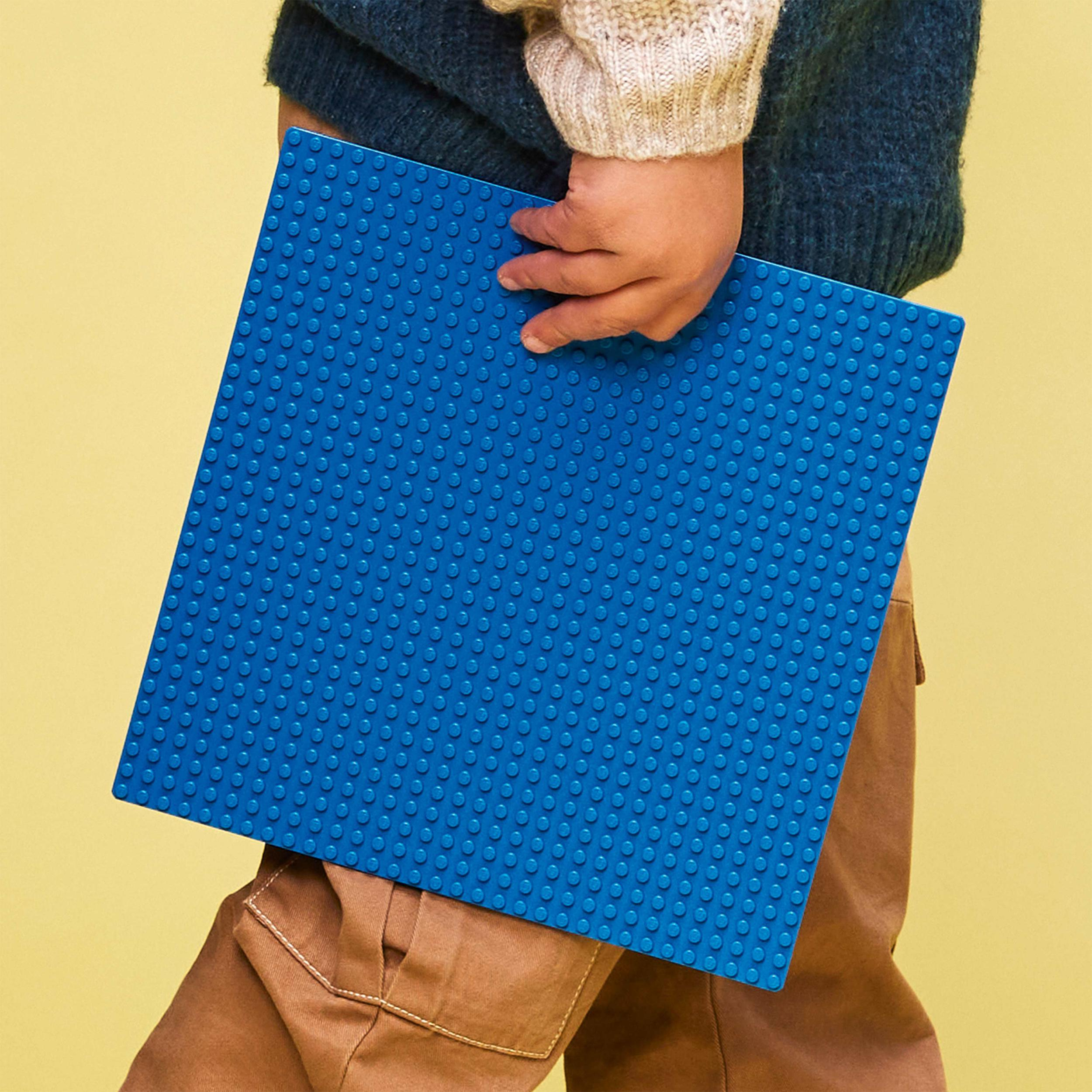 Ein Kind in einem blauen Pullover und braunen Hosen hält eine große blaue LEGO-Grundplatte.