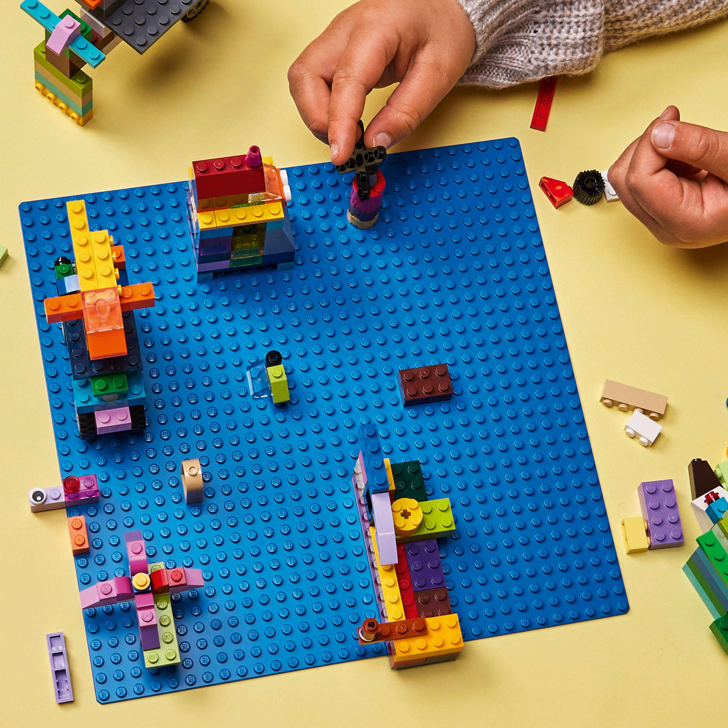 Hände bauen mit bunten Lego-Steinen auf einer blauen Grundplatte.