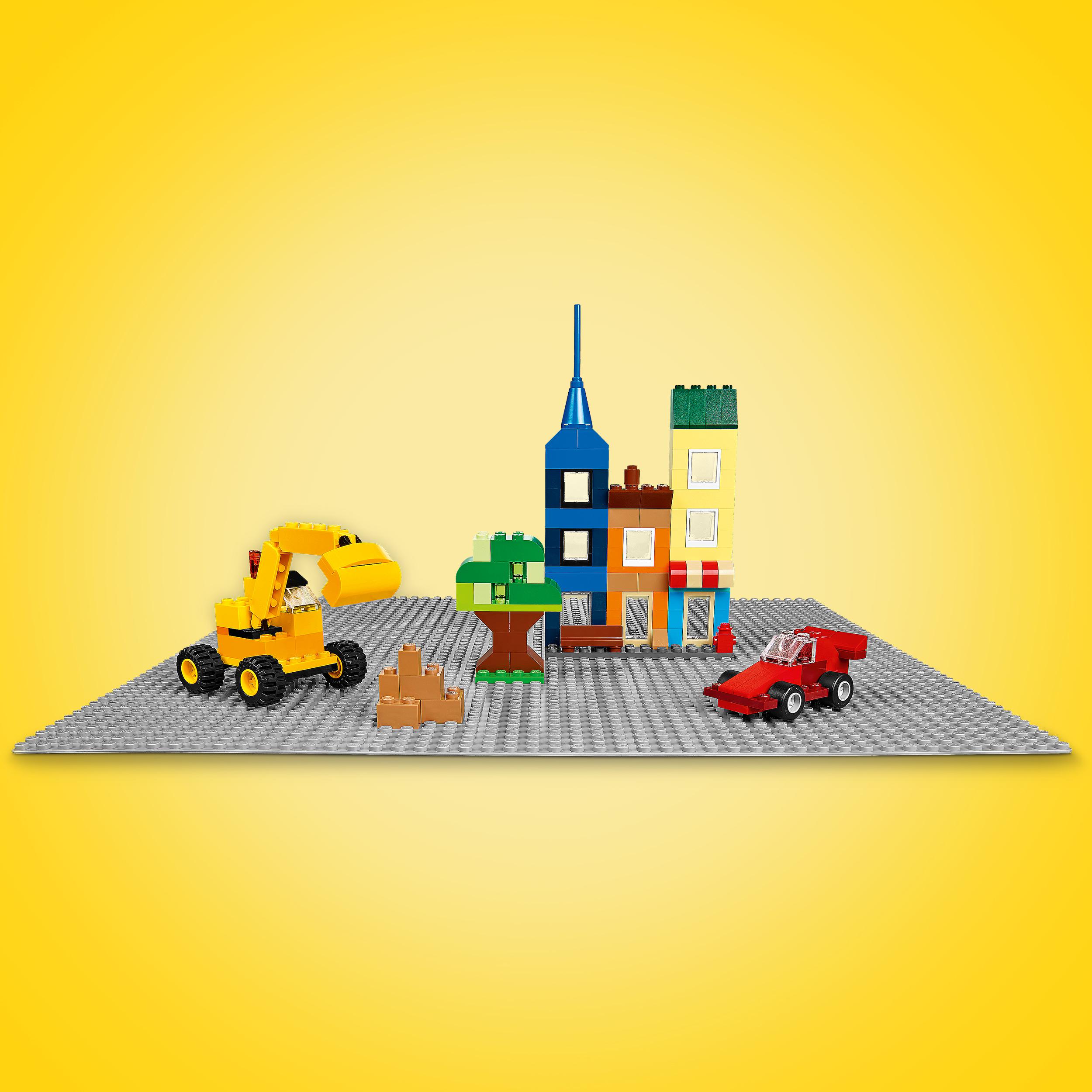 Eine Lego-Szene auf einer grauen Grundplatte mit einem gelben Bagger, einem roten Auto, Gebäuden und einem Baum.