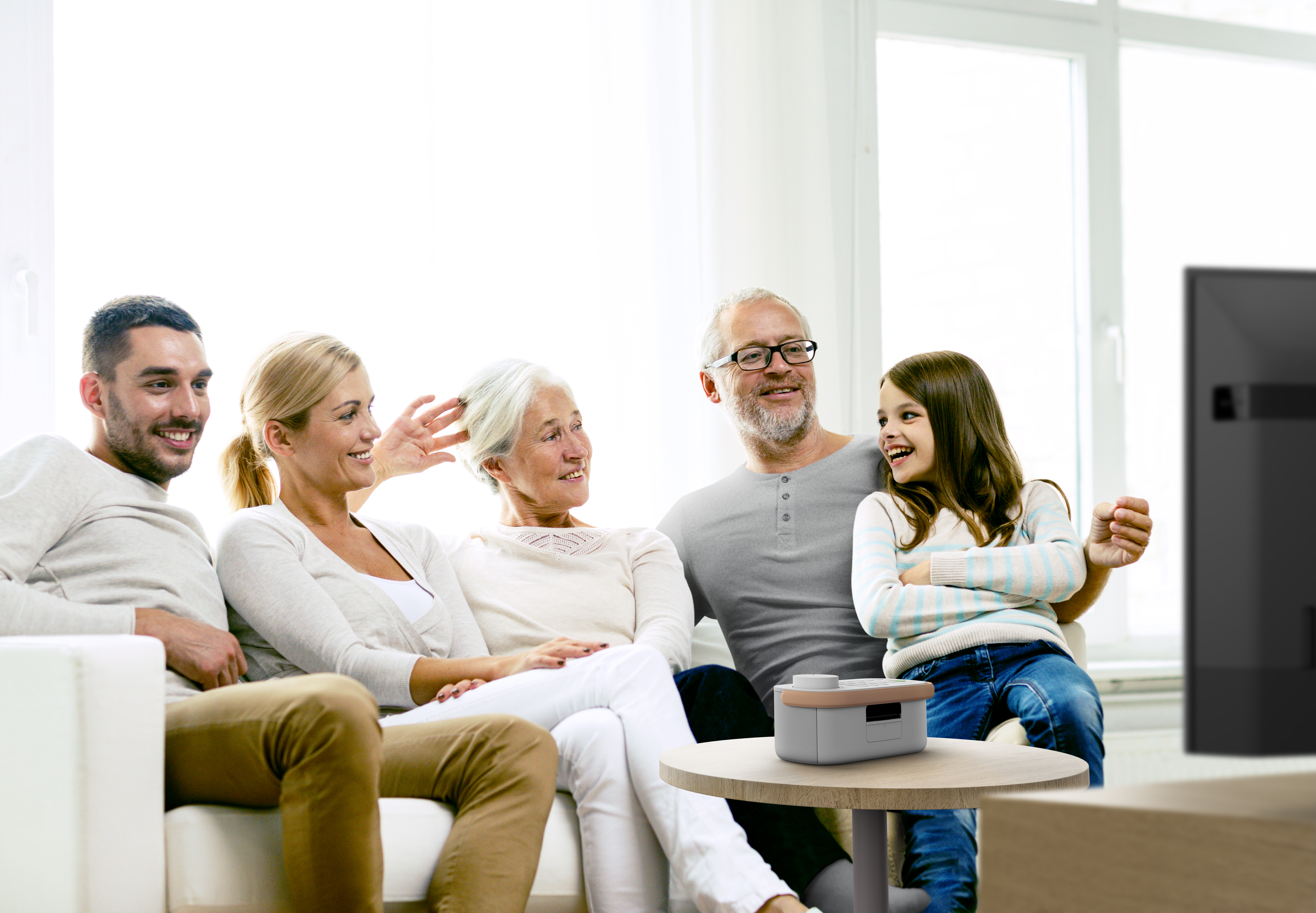 Una familia de cinco personas está sentada en un sofá, mirando algo fuera de cuadro. Una mesa pequeña con un dispositivo.