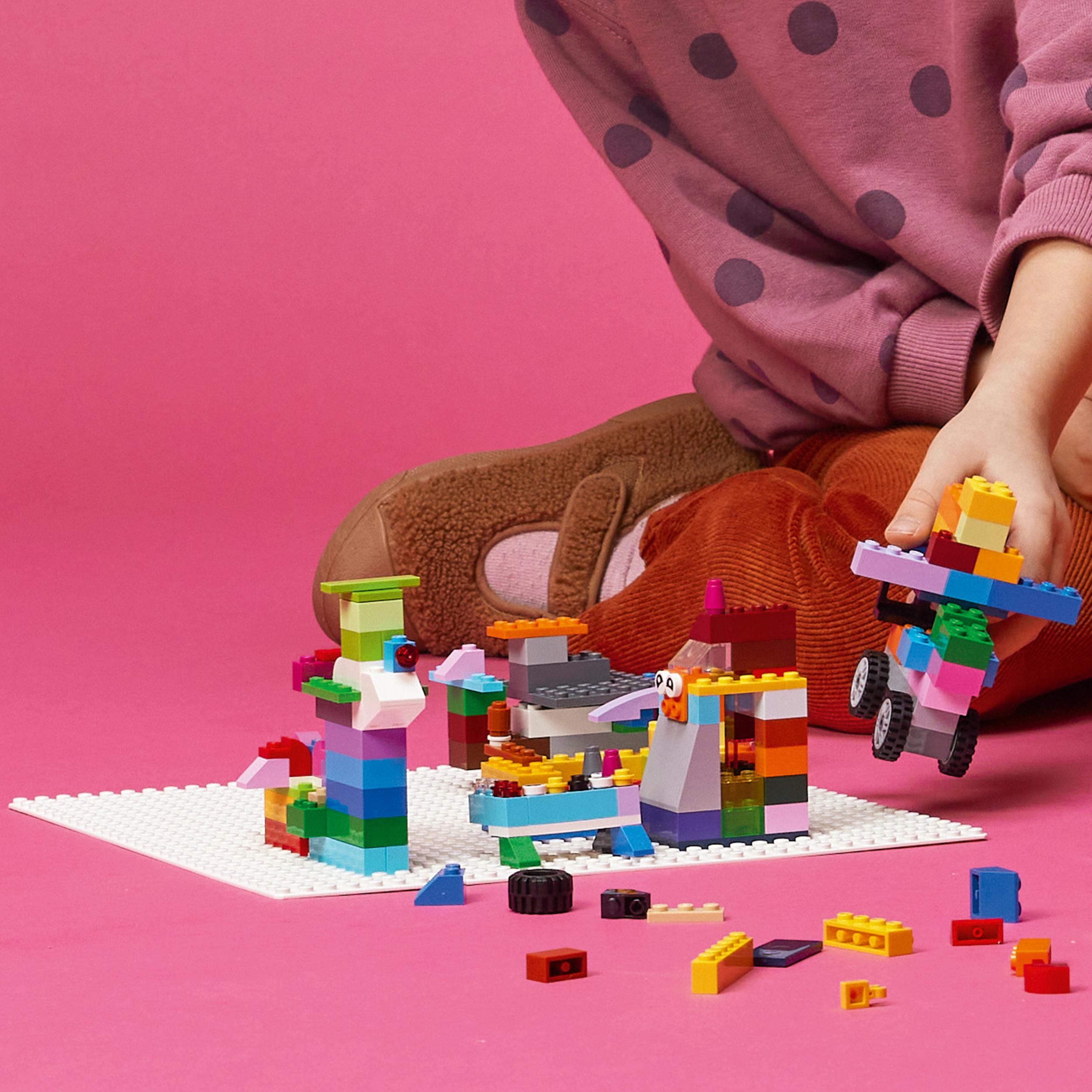 Ein Kind spielt mit bunten Lego-Steinen auf einer weißen Grundplatte vor rosa Hintergrund.