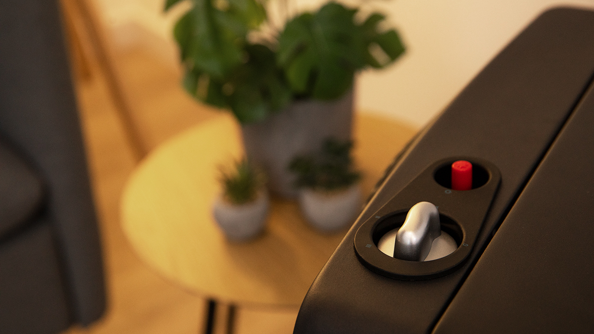 Dispositivo negro con botón rojo y perilla plateada. Plantas y una mesa en el fondo borroso.