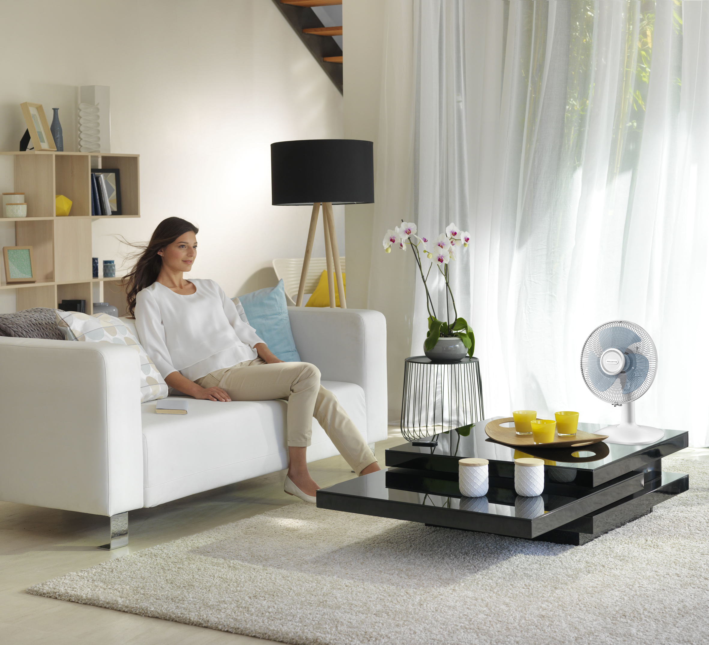 Mujer en sofá blanco en salón con mesa negra y ventilador.